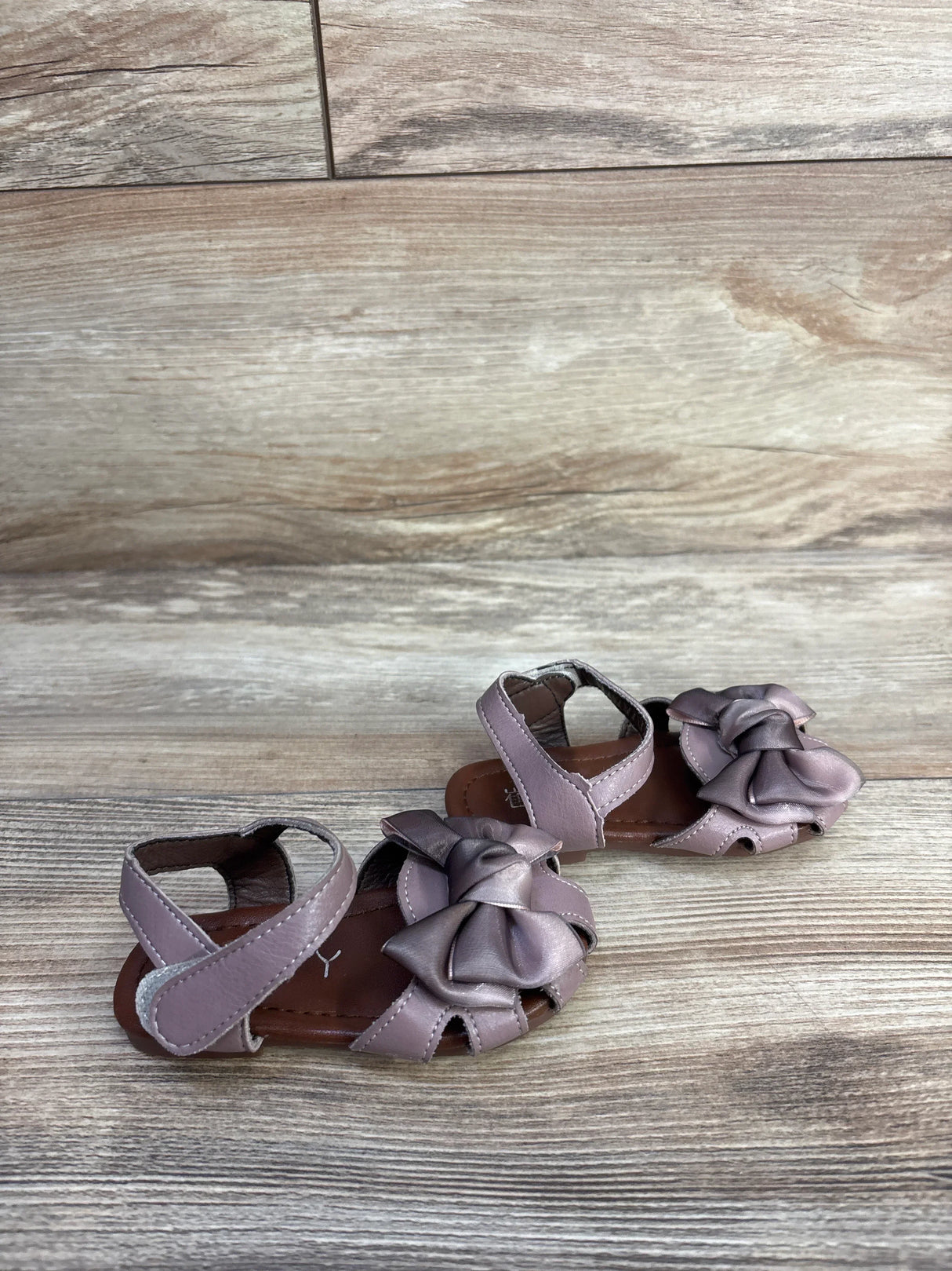 Front three-quarter view of EYY mauve baby sandals showing large bow detail and Velcro ankle straps on wood background