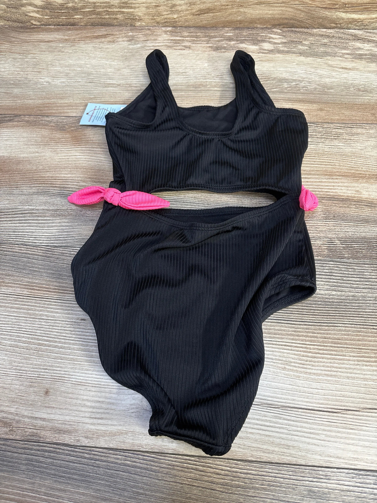 Flat-lay of black ribbed girls' one-piece cut-out swimsuit showcasing pink side tie accents on wood background