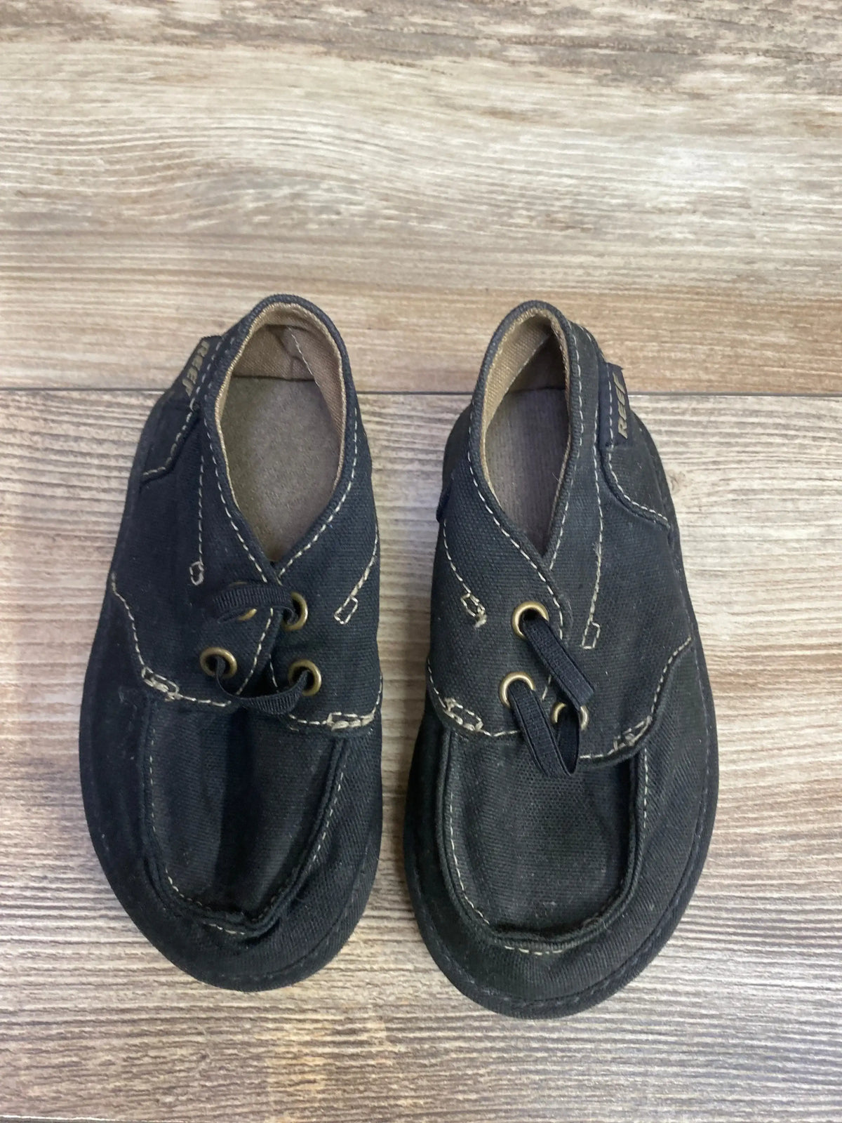 Top view of Reef Deckhand 3 toddler black canvas sneakers with beige contrast stitching on wood floor