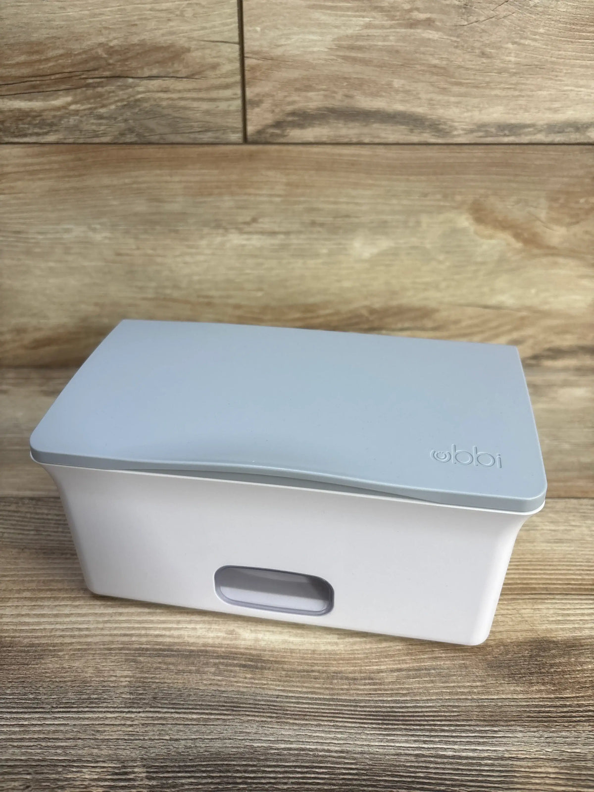 Front three-quarter view of Ubbi wipes dispenser showcasing its grey lid and front dispensing opening on a wood surface