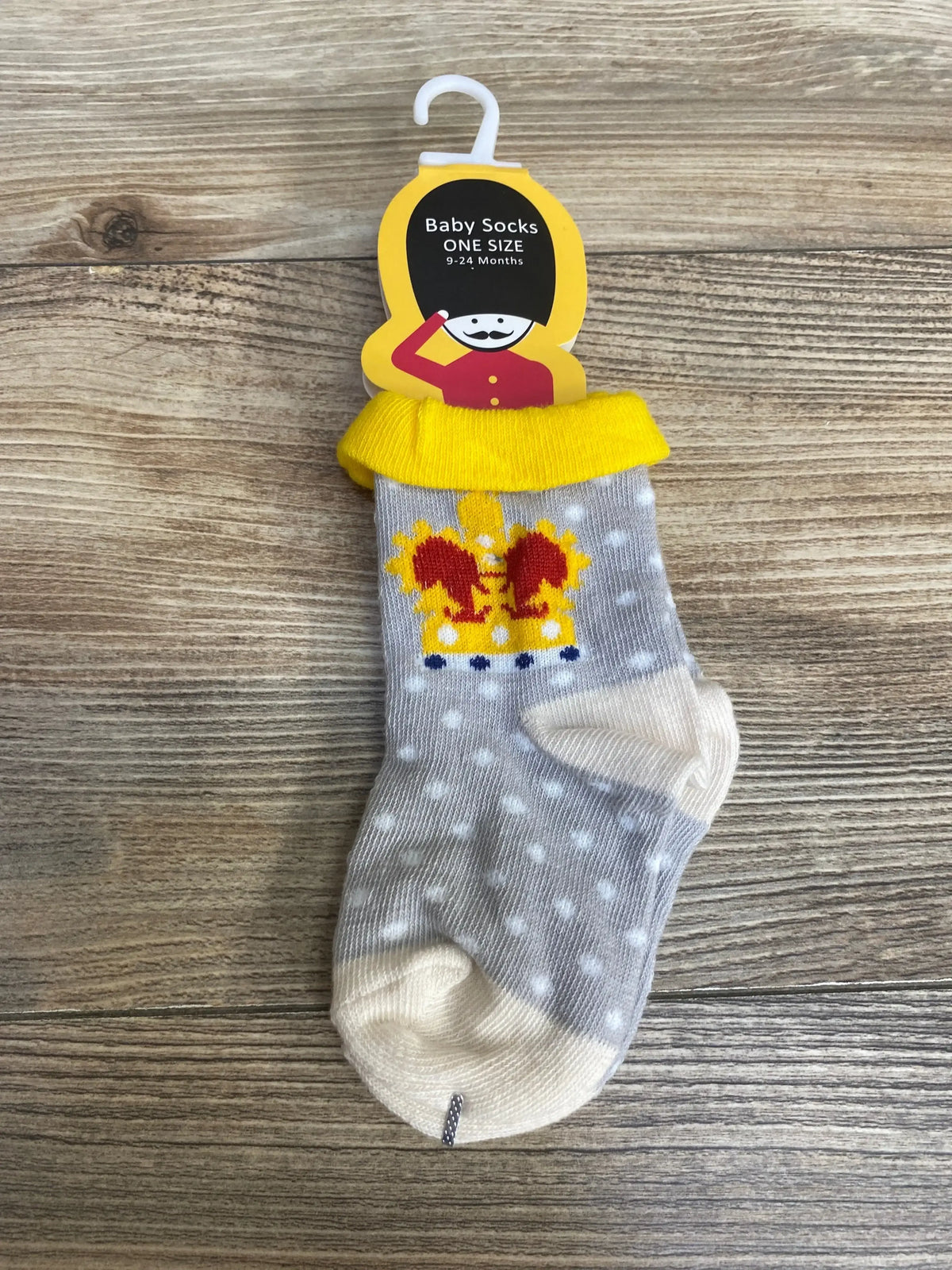 Top-down view of grey polka-dot baby sock with yellow cuff and embroidered crown, one size 9-24 months, on wood background