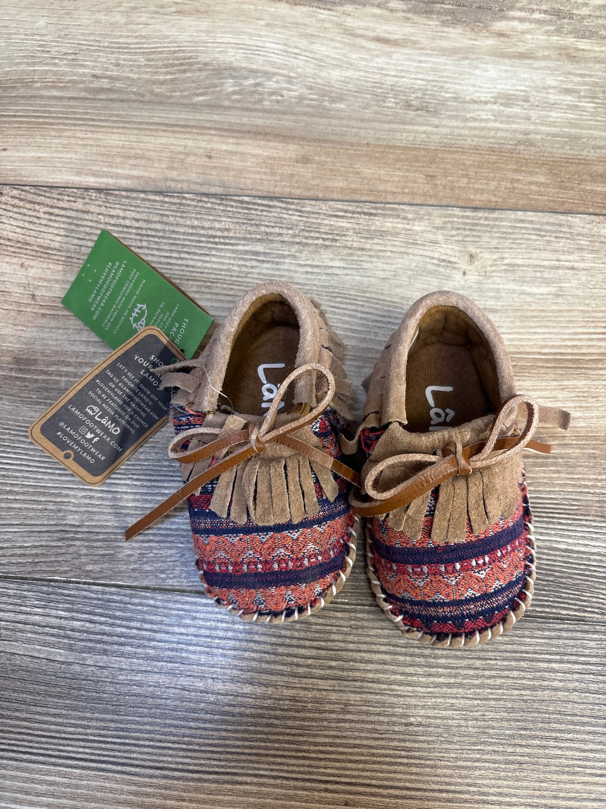 Brown and red baby moccasins with fringe detail and patterned fabric, on wood floor