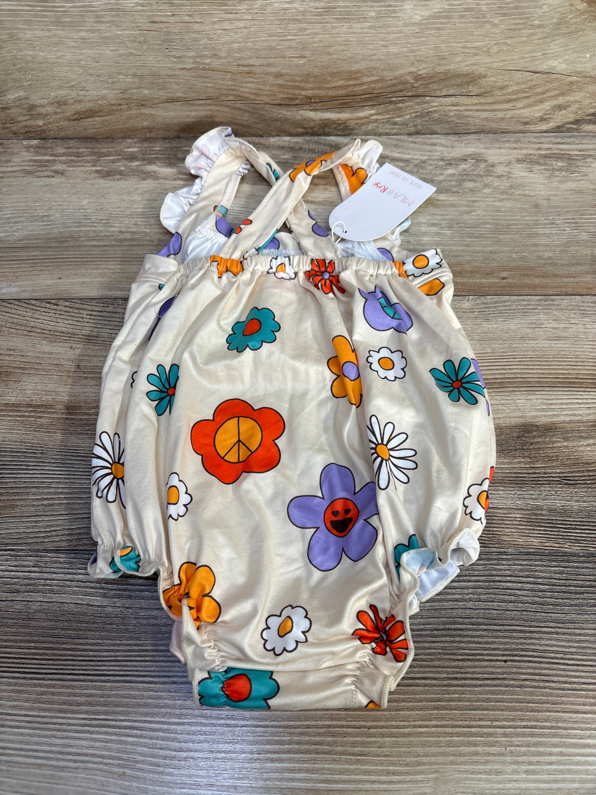 Flat lay of beige floral crossback baby romper showing crisscross straps, elastic leg openings and colorful flower print on wood