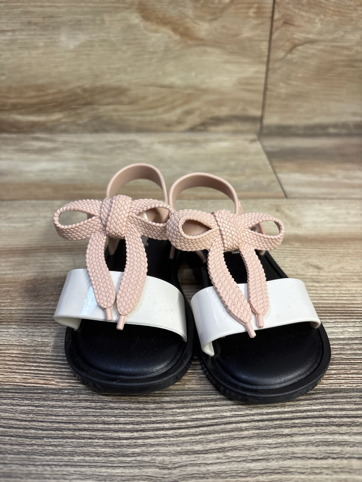 Front view of Mini Melissa Mar toddler sandals with pink braided bow, white toe strap and black footbed on wood background
