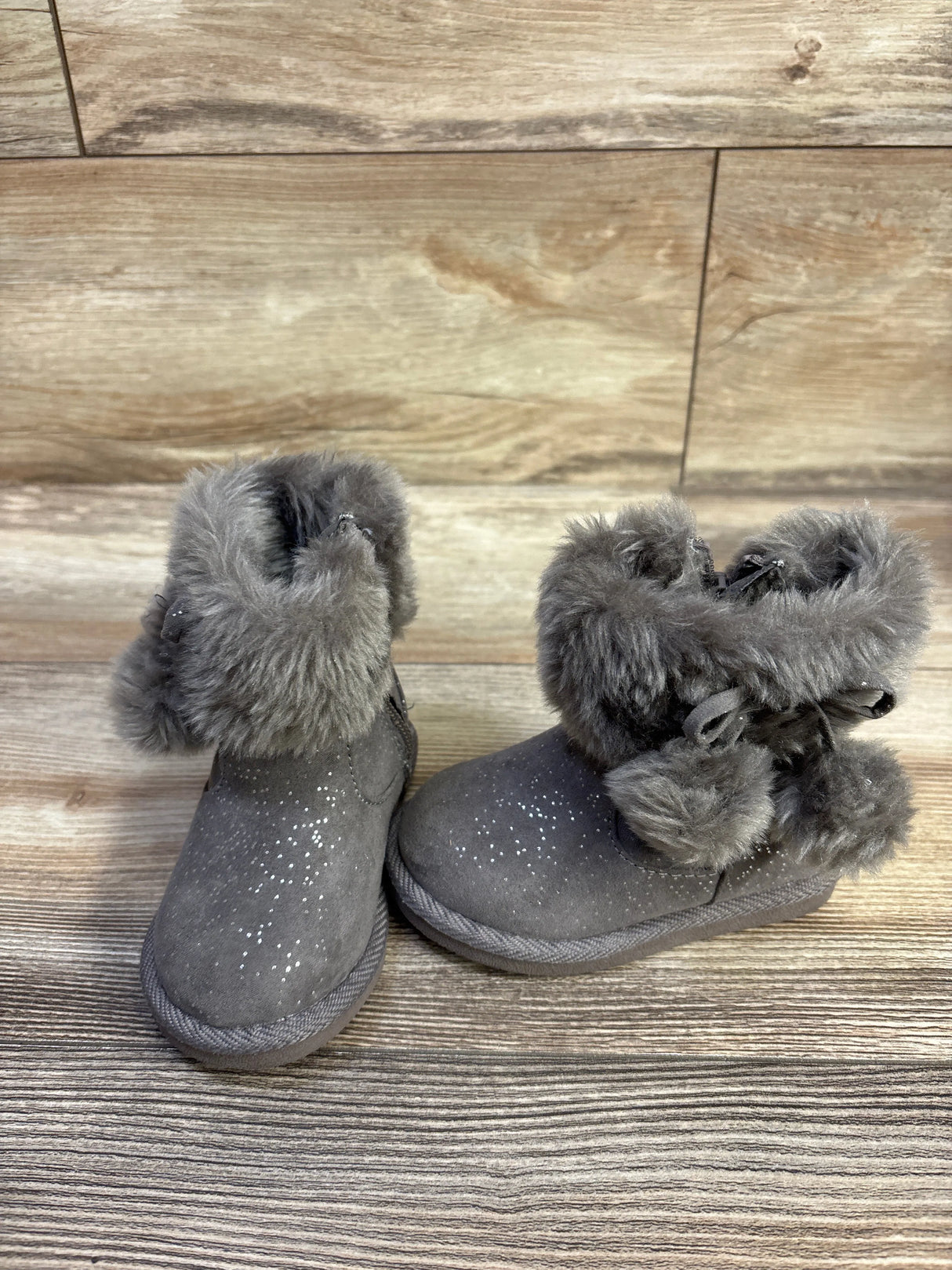Three-quarter view of Jumping Beans Coco Easy Zip boots in grey with faux-fur cuff, pom-poms and glitter on wood
