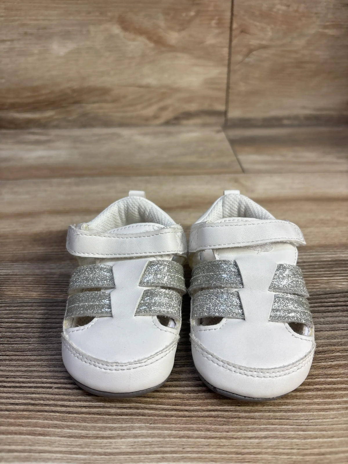 Front view of Surprize by Stride Rite fisherman sandals showcasing white uppers and silver glitter straps with a wood grain background