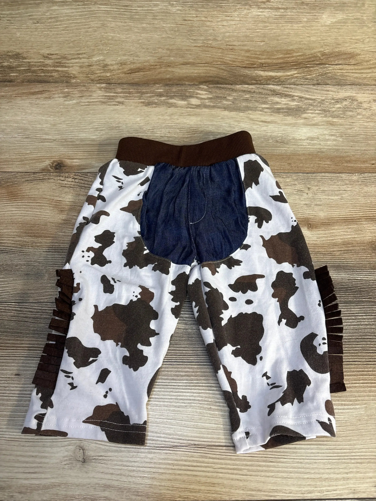 Cow print kids pants with denim back patch and brown ruffles, laid on wood floor