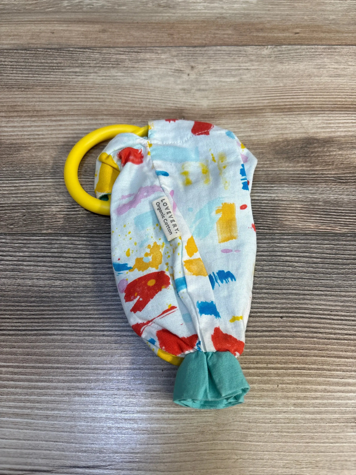Top-down view of Lovevery Organic Cotton Sensory Pouch with paint-splatter fabric, yellow teething ring, green knotted end, on wood surface