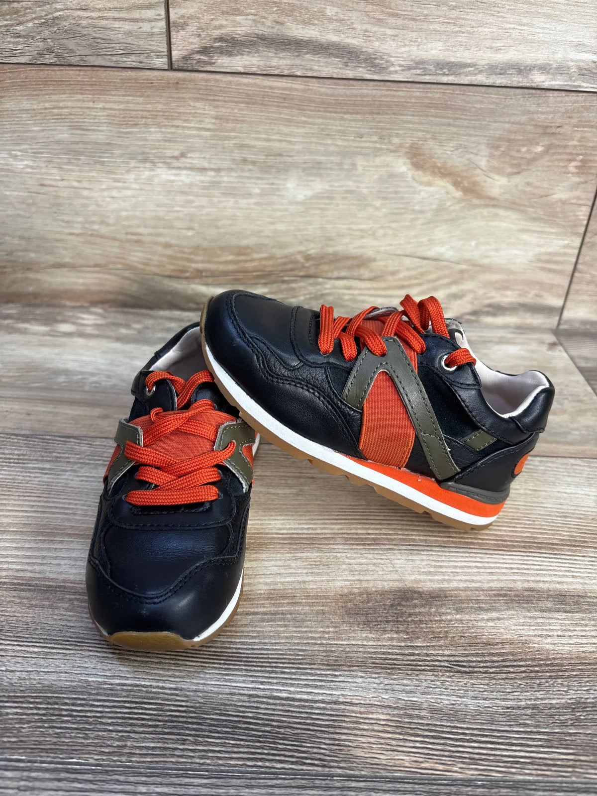 Angled view of black Dolce & Gabbana Bambino leather sneakers with orange laces, olive panels, and gum soles on a wood floor