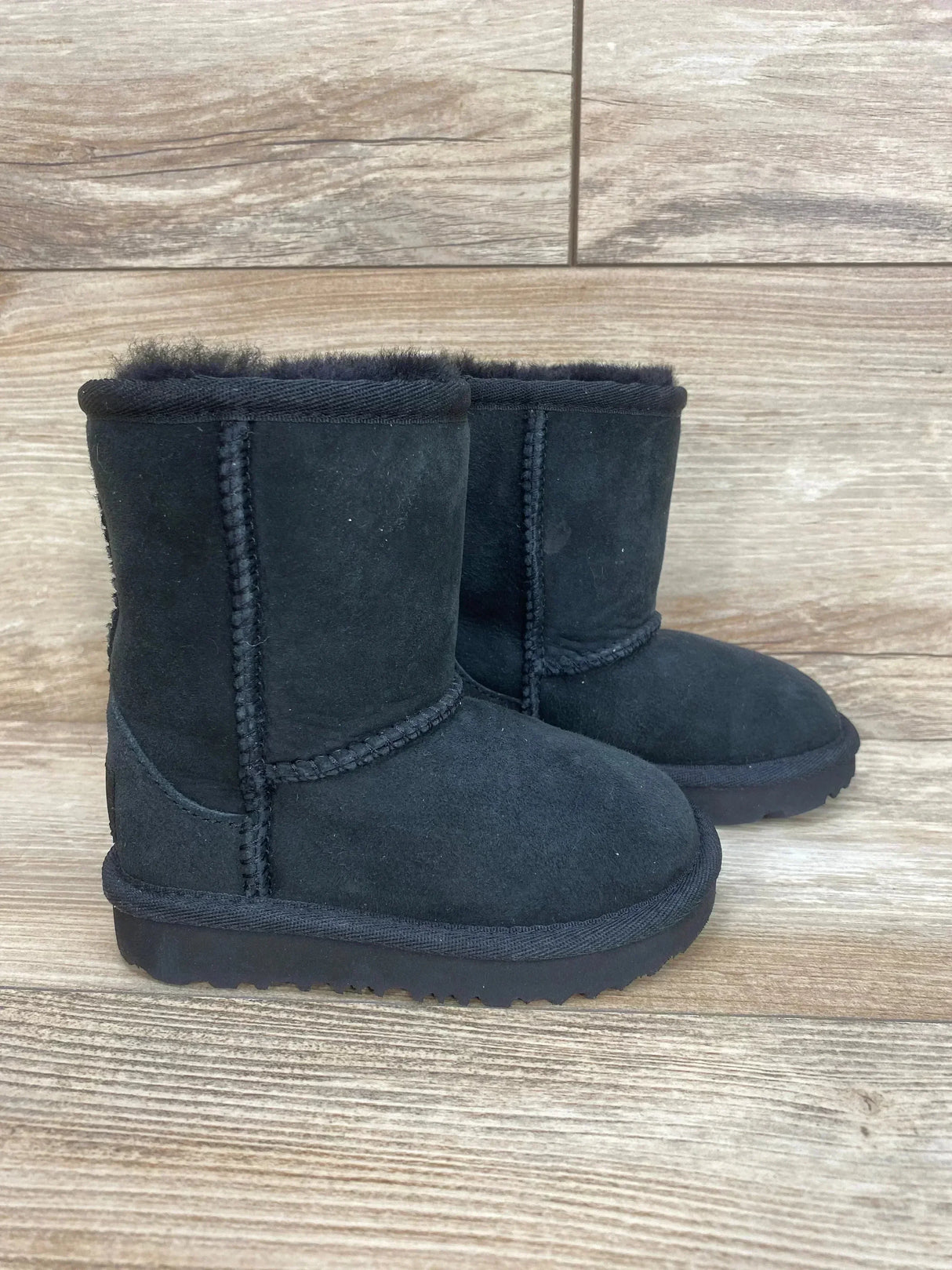 Side view of UGG Toddlers' Classic II black suede toddler boot with sheepskin lining and stitched seams on wood floor, size 6C