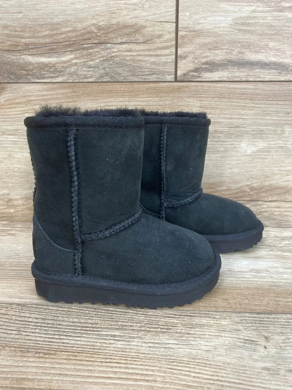 Side view of UGG Toddlers' Classic II black suede toddler boot with sheepskin lining and stitched seams on wood floor, size 6C
