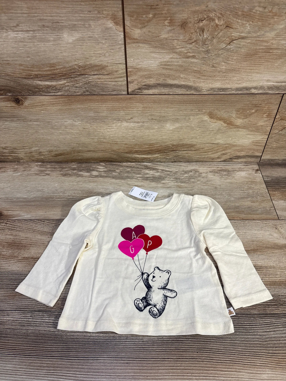 Front view of Baby Gap cream long puff sleeve shirt in size 6-12 months showcasing teddy bear and balloon graphic on a wood floor
