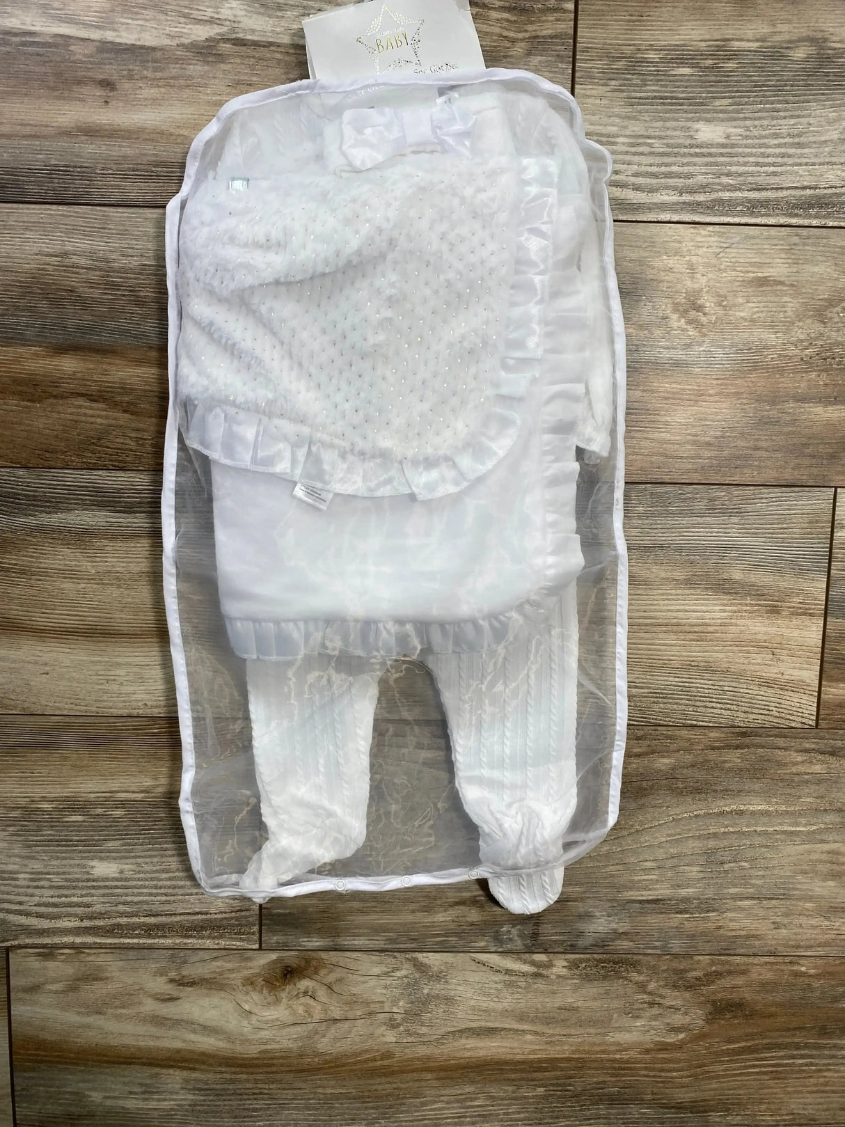 Top-down view of white 5-piece christening set size 6M showcasing ruffled satin-trim blanket and textured knit footed pants in a sheer storage bag on wood floor