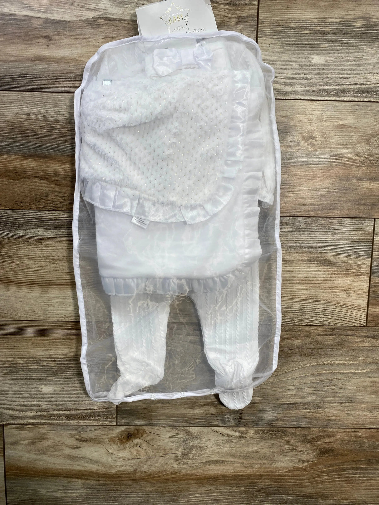 Top-down view of white 5-piece christening set size 6M showcasing ruffled satin-trim blanket and textured knit footed pants in a sheer storage bag on wood floor