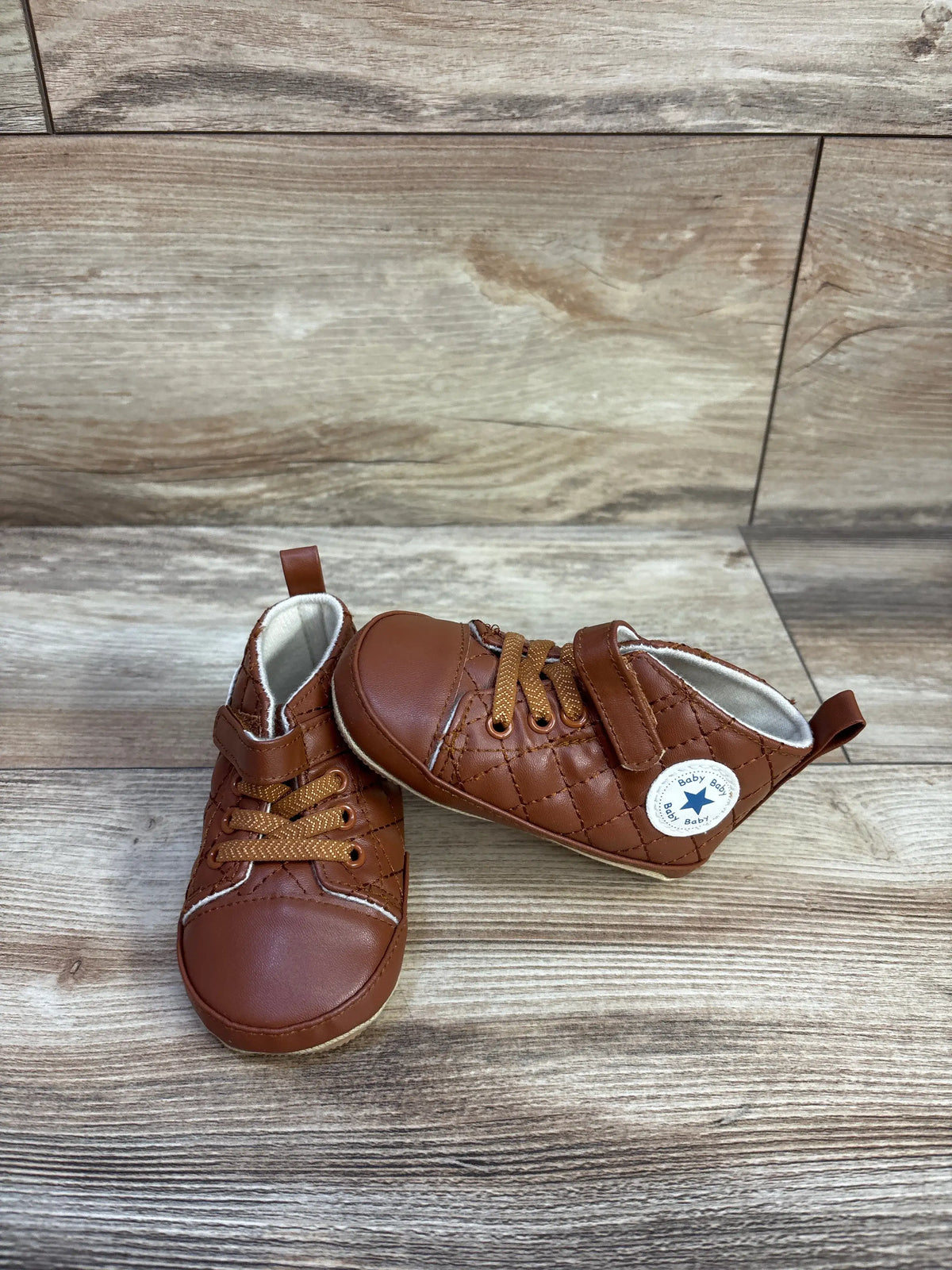 Front and side views of brown faux leather crib shoes showcasing quilted uppers and laces on a wood-look background
