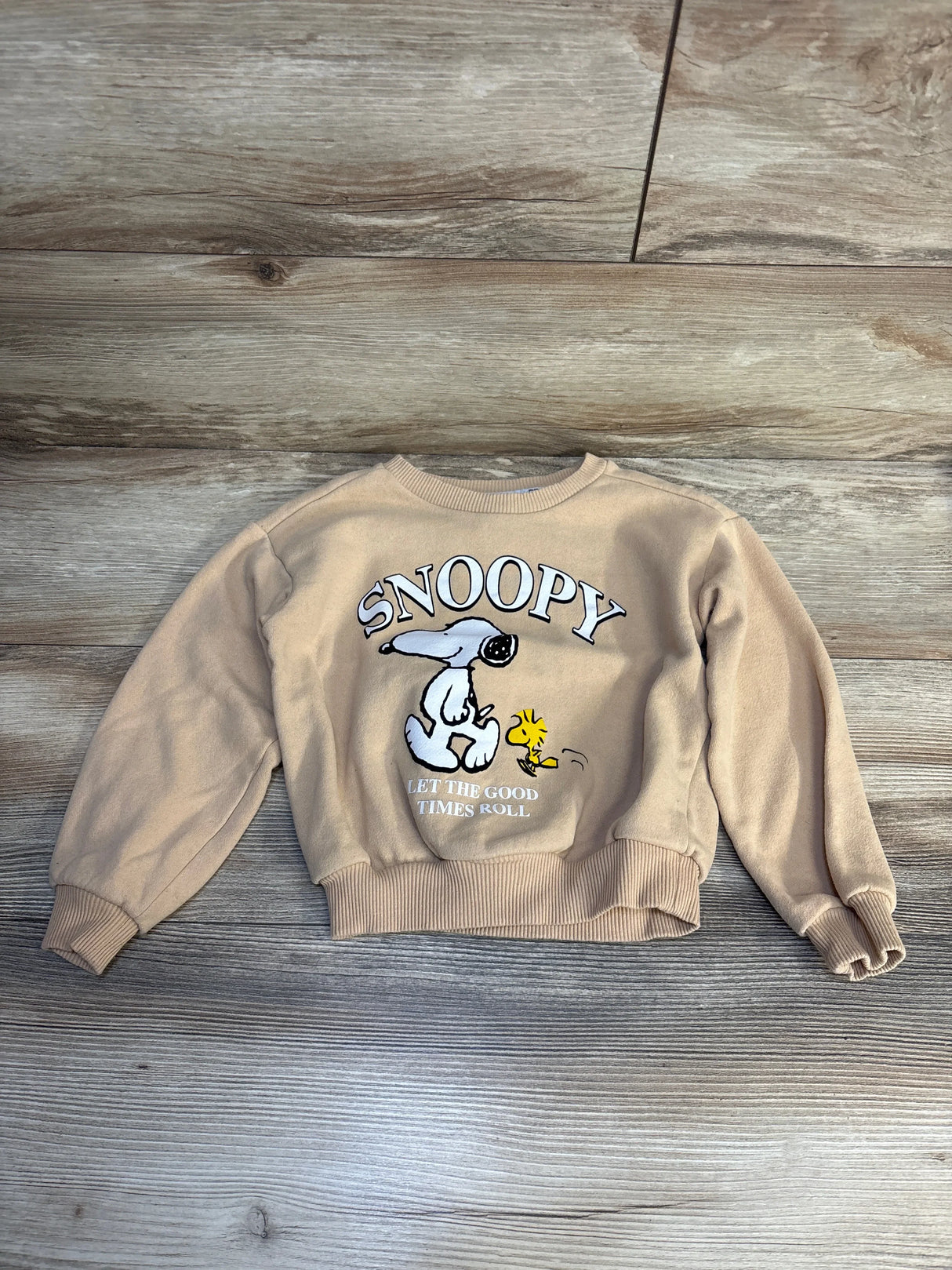 Tan kids Snoopy sweatshirt with Woodstock, cartoon print, long sleeves, laid on wood floor