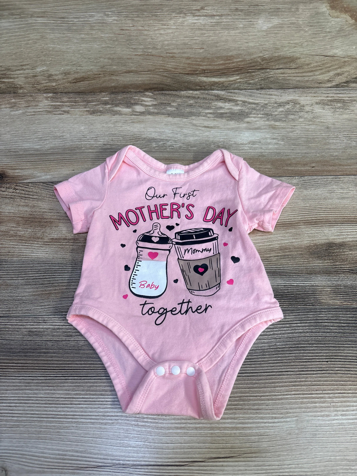 Flat-lay of pink 0-3M bodysuit showing 'Our First Mother's Day' print with baby bottle and coffee cup on wood background