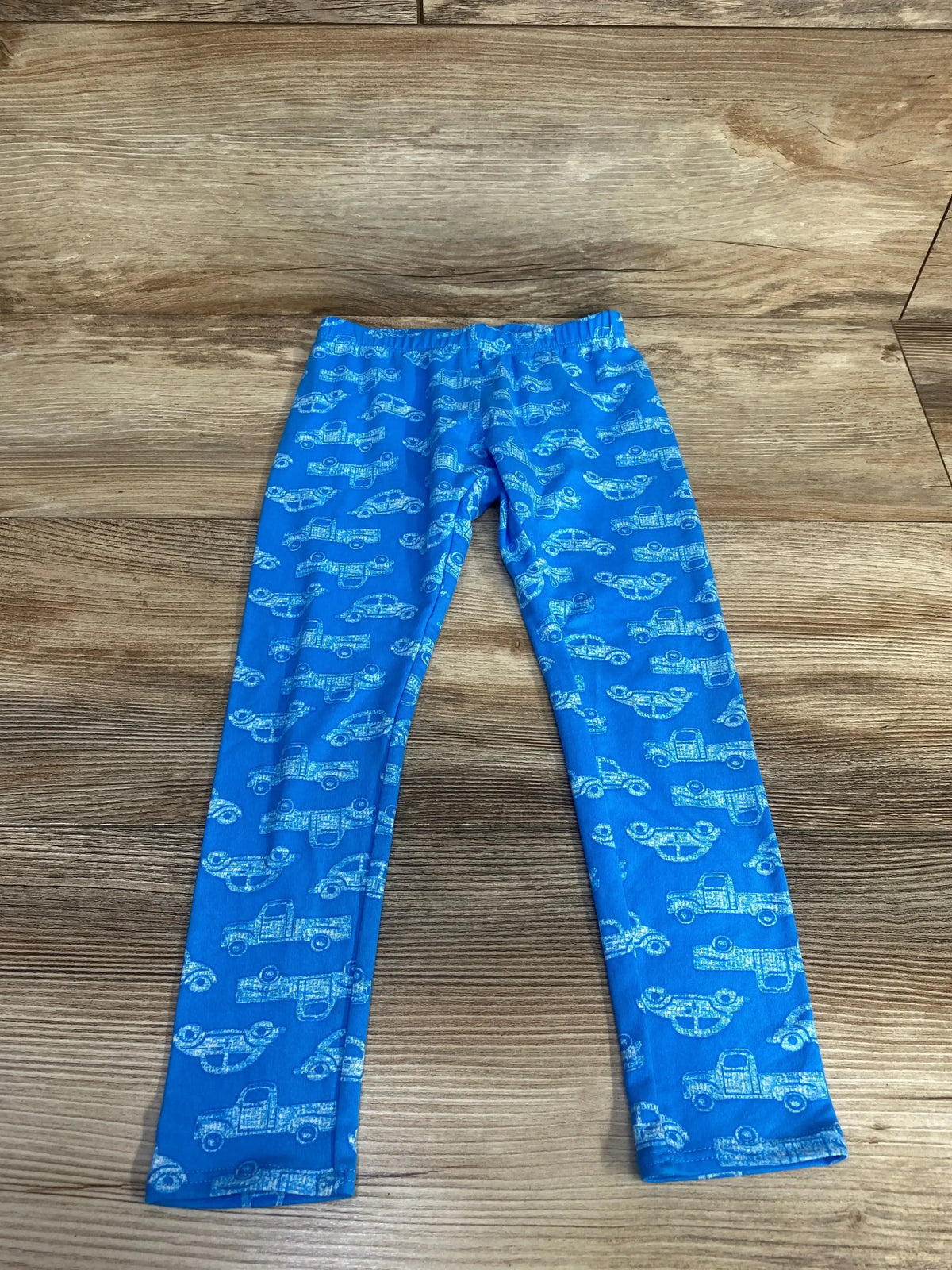 Flat lay of blue toddler leggings (size 3-4T) showcasing white car print and elastic waistband on wood floor background