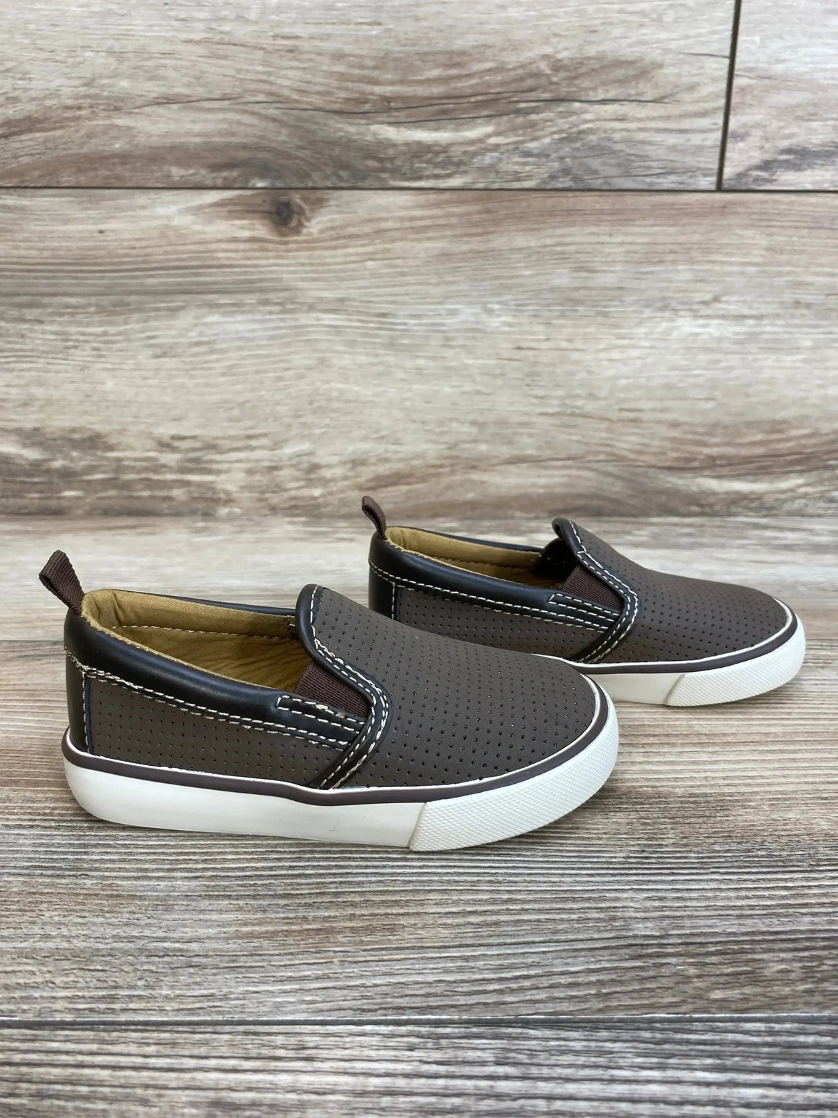Side view of brown toddler slip-on sneakers with perforated upper, white rubber sole, elastic sides, on wood background