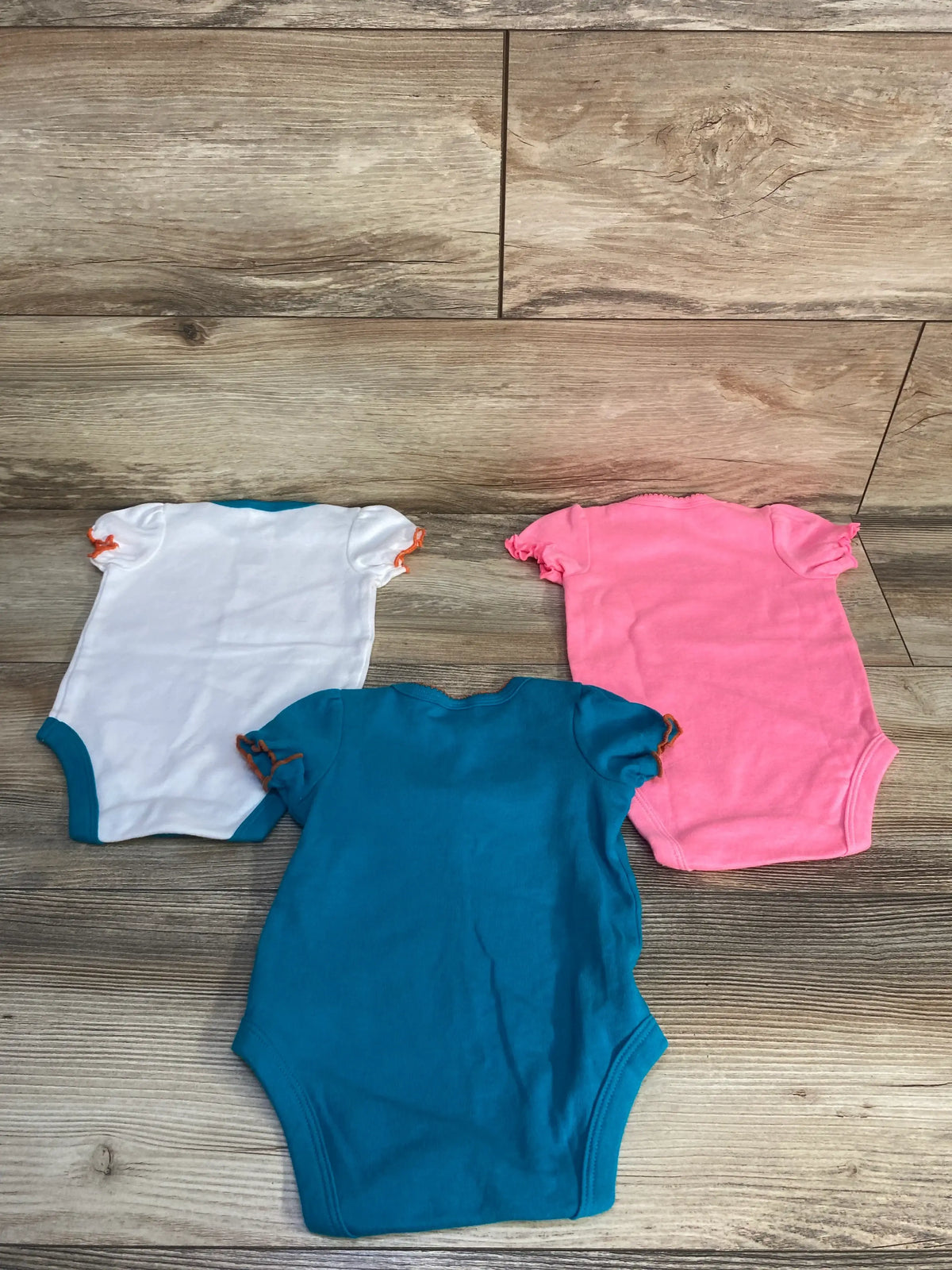 Flat lay of Miami Dolphins three pack bodysuits in white, pink, and teal with short sleeves on wood grain flooring