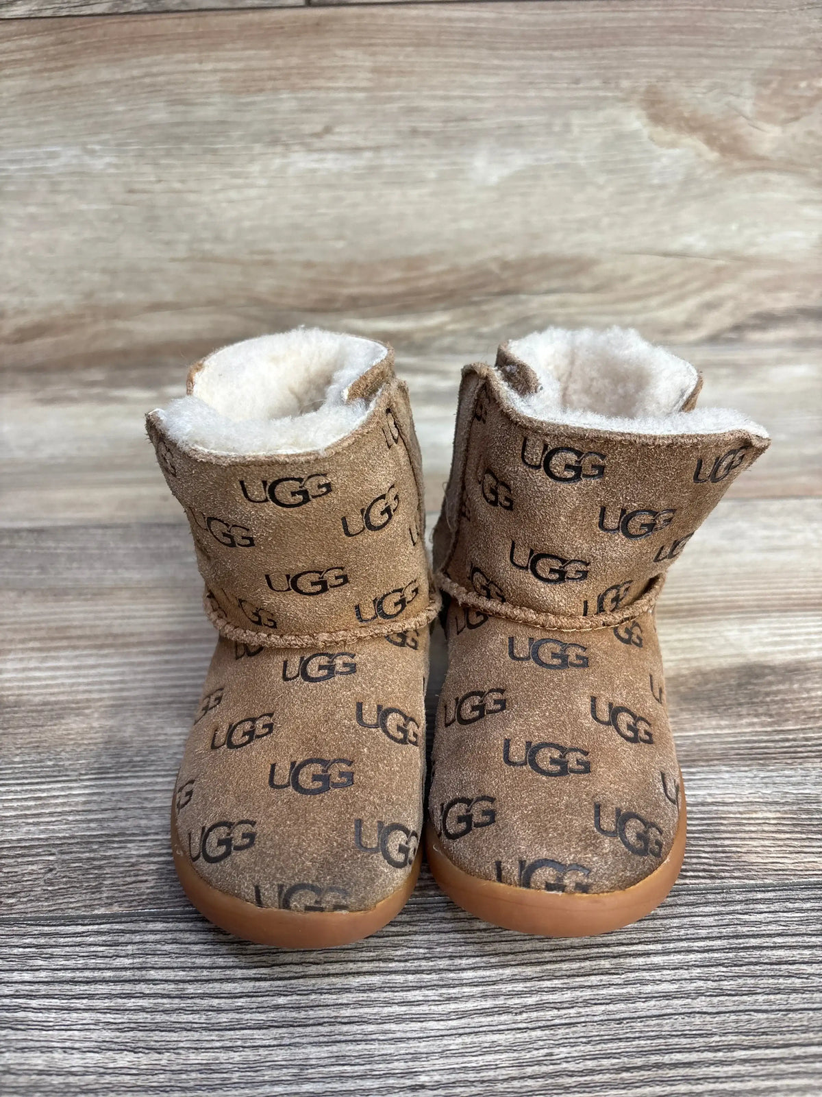 Pair of brown UGG baby boots with black UGG logos and white fleece lining on wooden floor