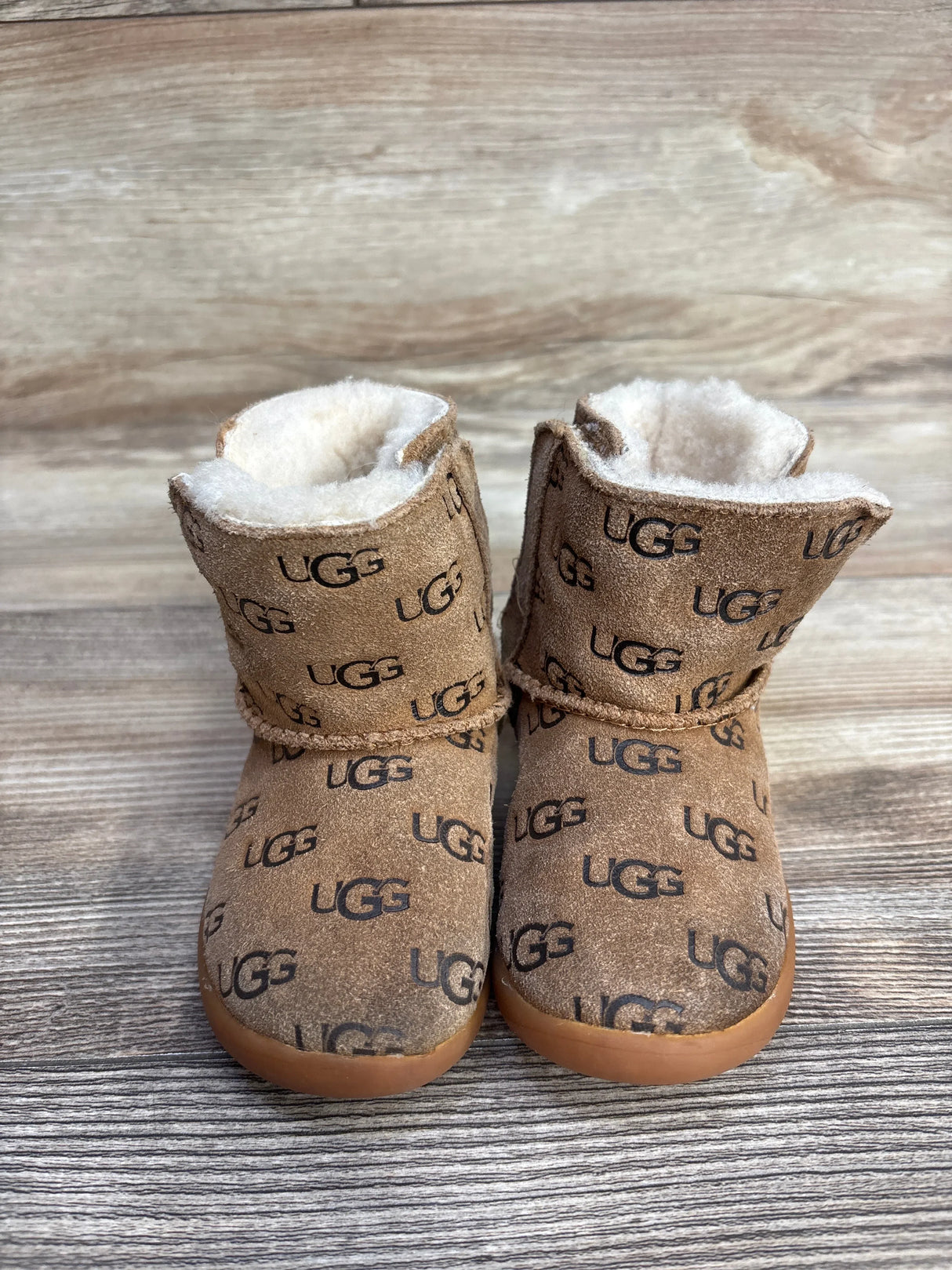 Pair of brown UGG baby boots with black UGG logos and white fleece lining on wooden floor