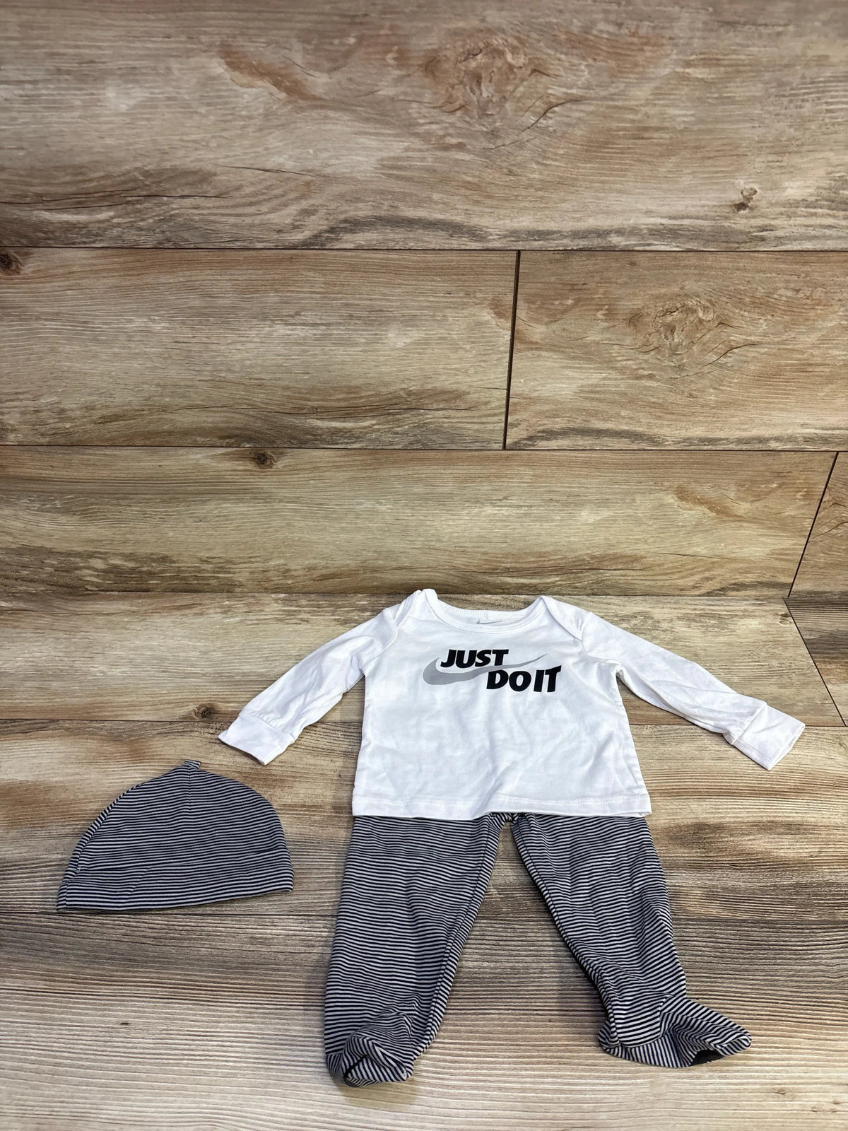 Flat lay of Nike 3-piece baby set with white long-sleeve JUST DO IT shirt, gray striped footed pants and matching hat on wood background