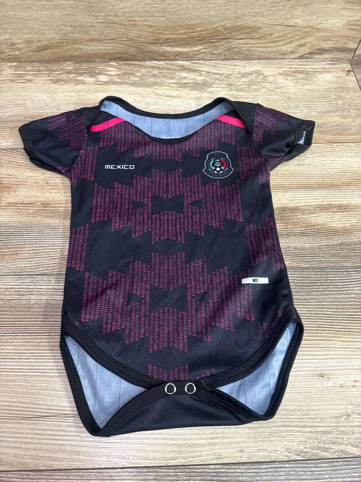 Front view of black Mexico soccer jersey bodysuit showcasing red geometric pattern, crest, and snap closures on wood flooring