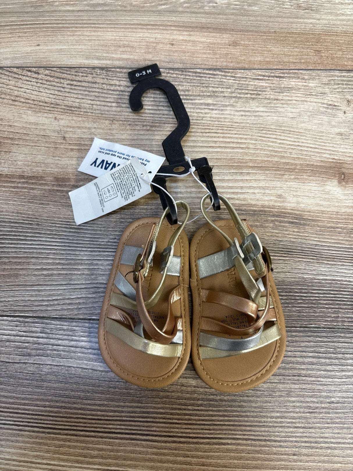 Infant metallic strappy sandals with tags, size 0-3 months, on wooden background