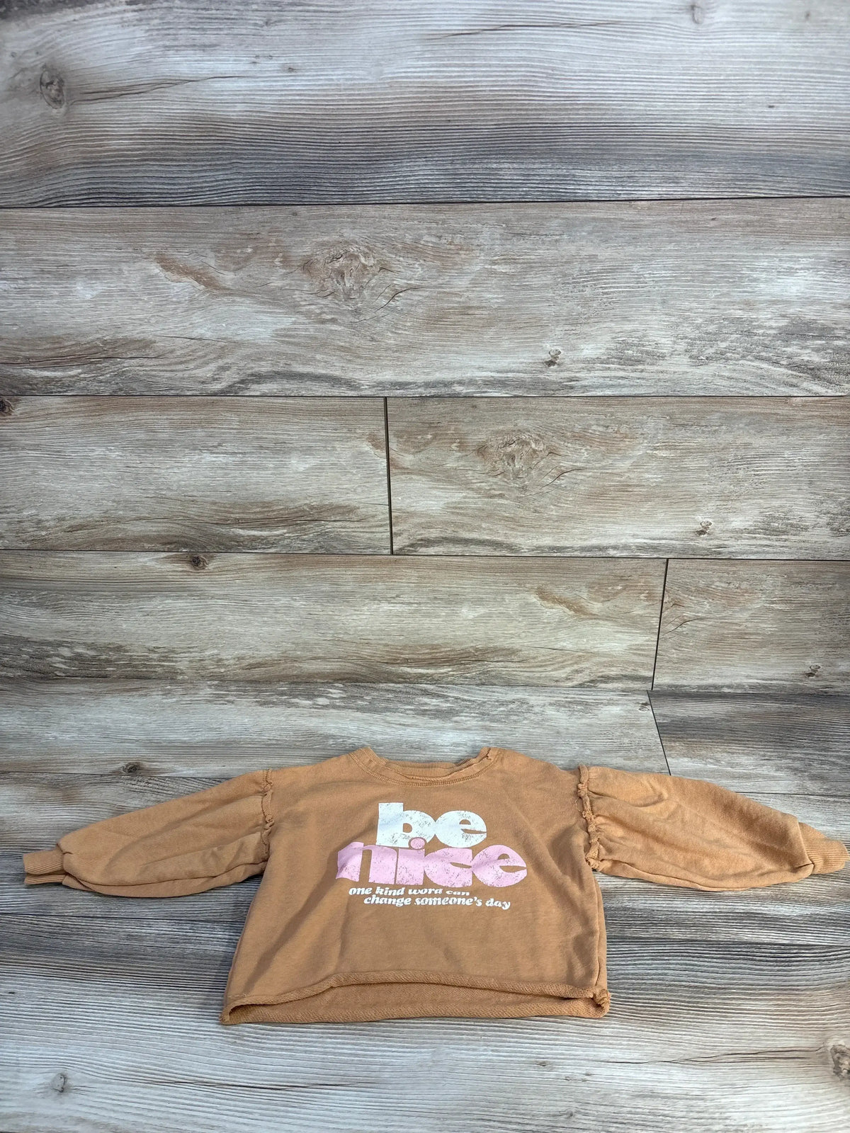 Tan kids' sweatshirt with 'be nice' text on wood background