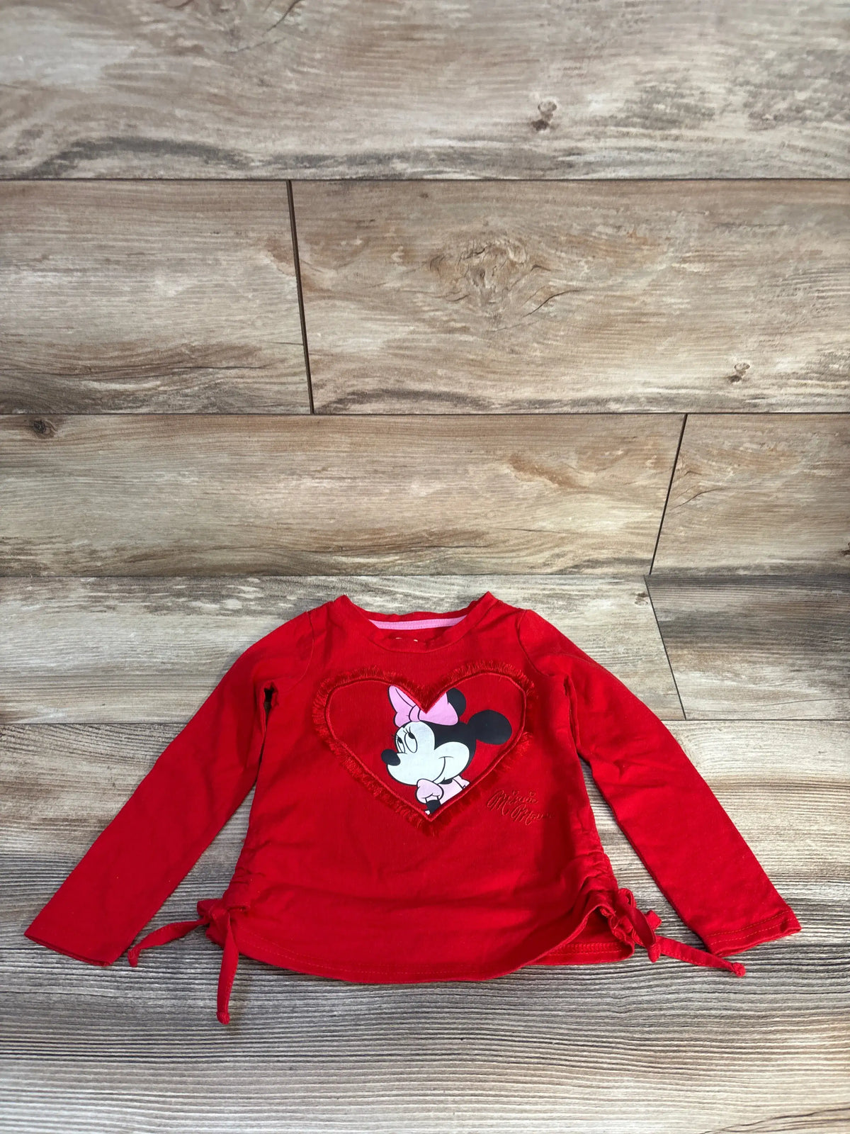 Front view of red Disney Minnie Mouse heart shirt showcasing long sleeves, heart graphic, and side ties on wood background
