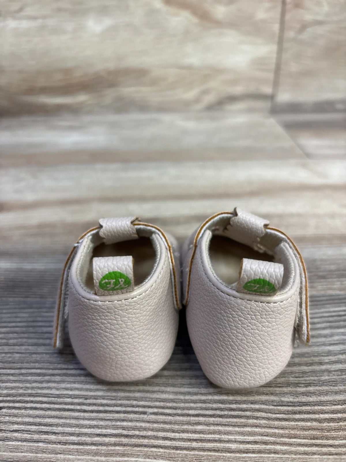 Rear view of First Steps soft sole shoes in beige showing textured leather, heel loops and Velcro straps on wood backdrop