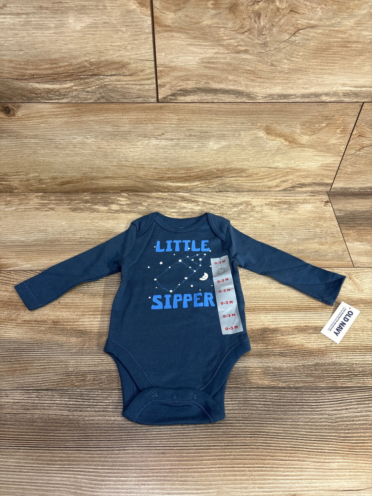 Flat lay of navy long-sleeve baby bodysuit (0-3M) showcasing ‘LITTLE SIPPER’ graphic, three crotch snaps and Old Navy tag on wood background