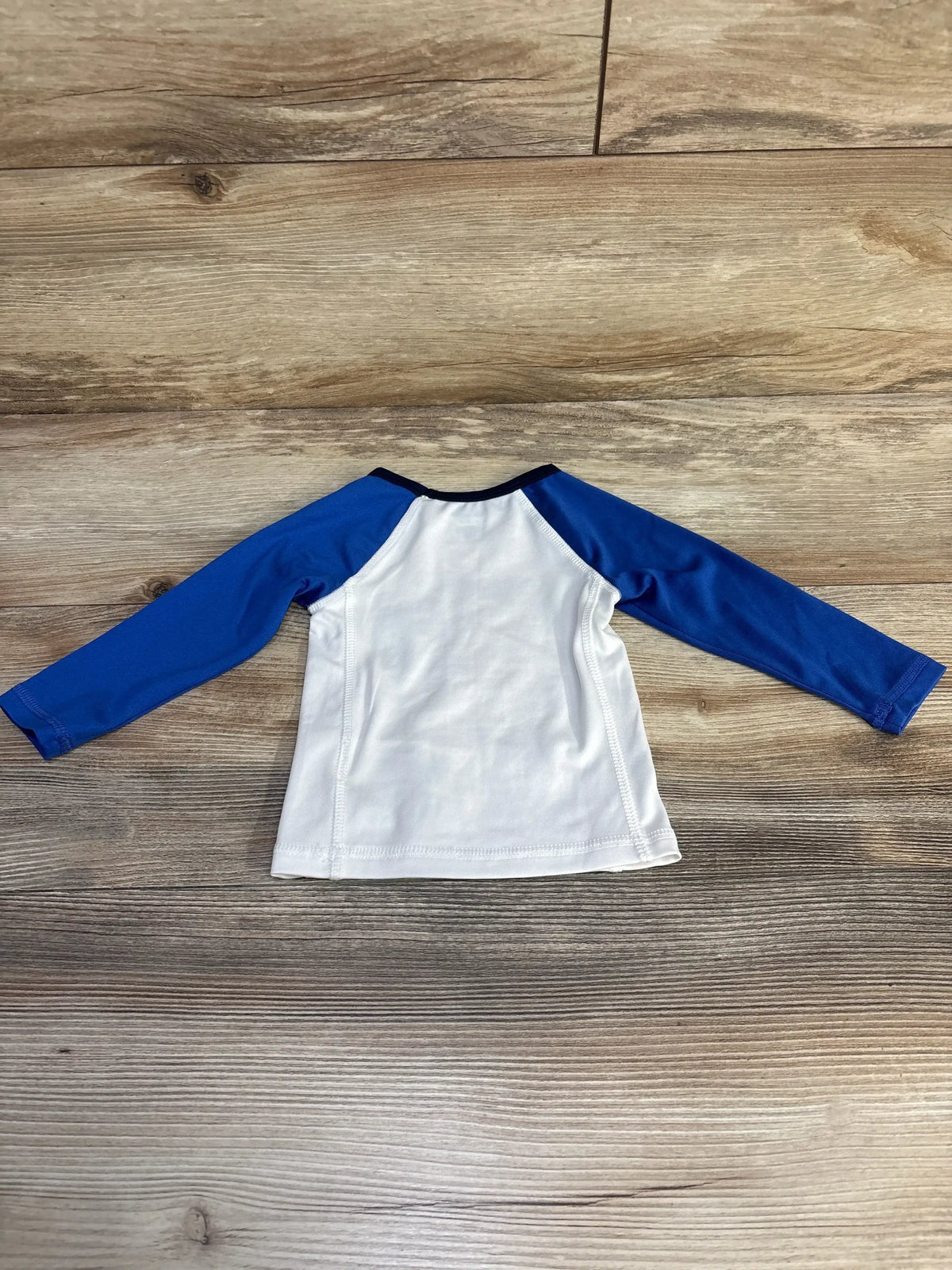 Back view of Ralph Lauren baby rash guard with white body blue raglan sleeves and navy neckline on wood background