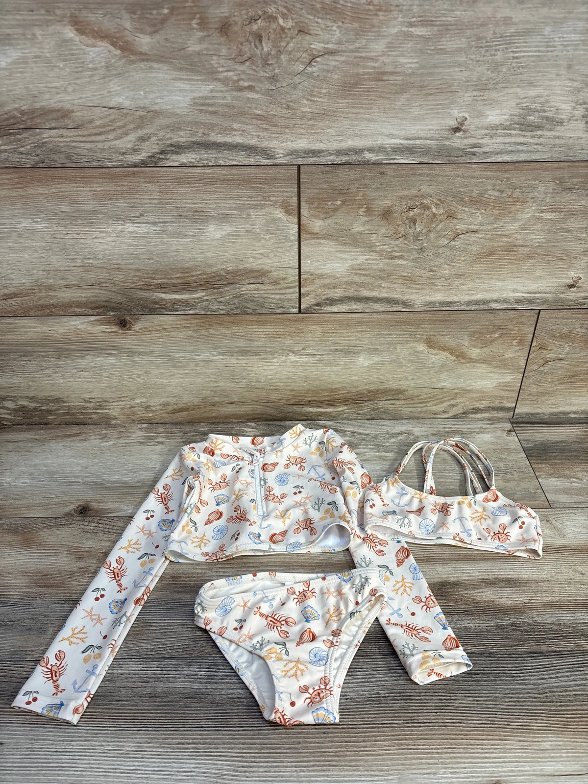 Flat lay of Rylee and Cru 3-piece cropped rashguard set showcasing nautical ivory print and front zipper on wood