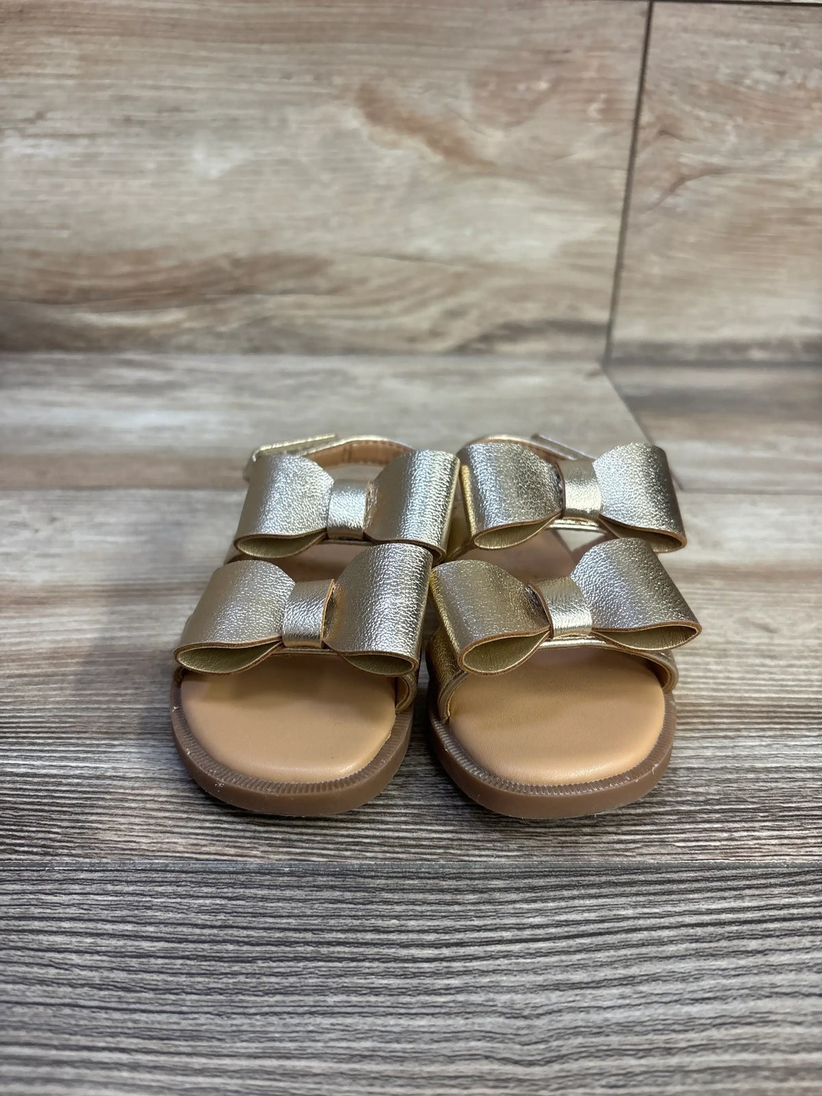 Front view of Cat & Jack Ruby Bow Sandals showcasing gold bow straps and open toes on a wood-grain background