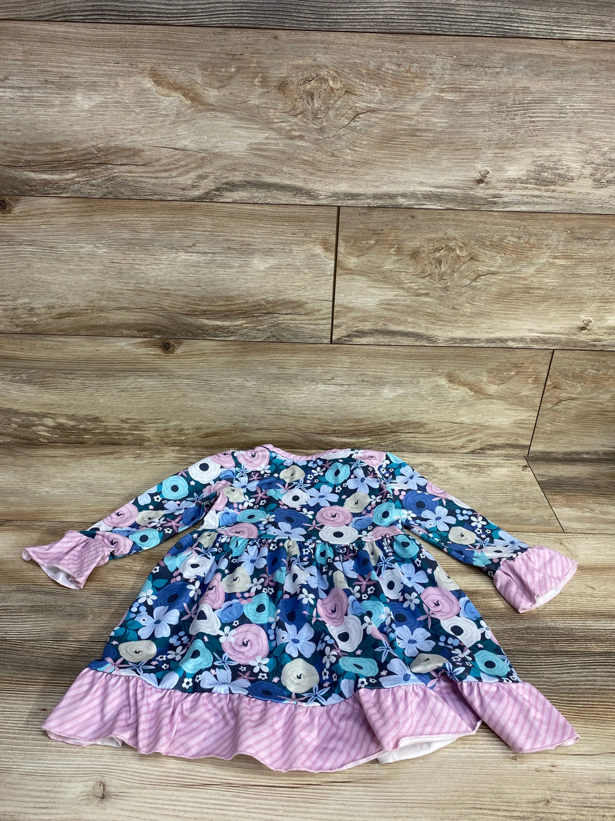 Flat lay of Pete + Lucy blue floral long sleeve toddler dress showcasing pink striped ruffle hem and sleeve cuffs on wood floor background