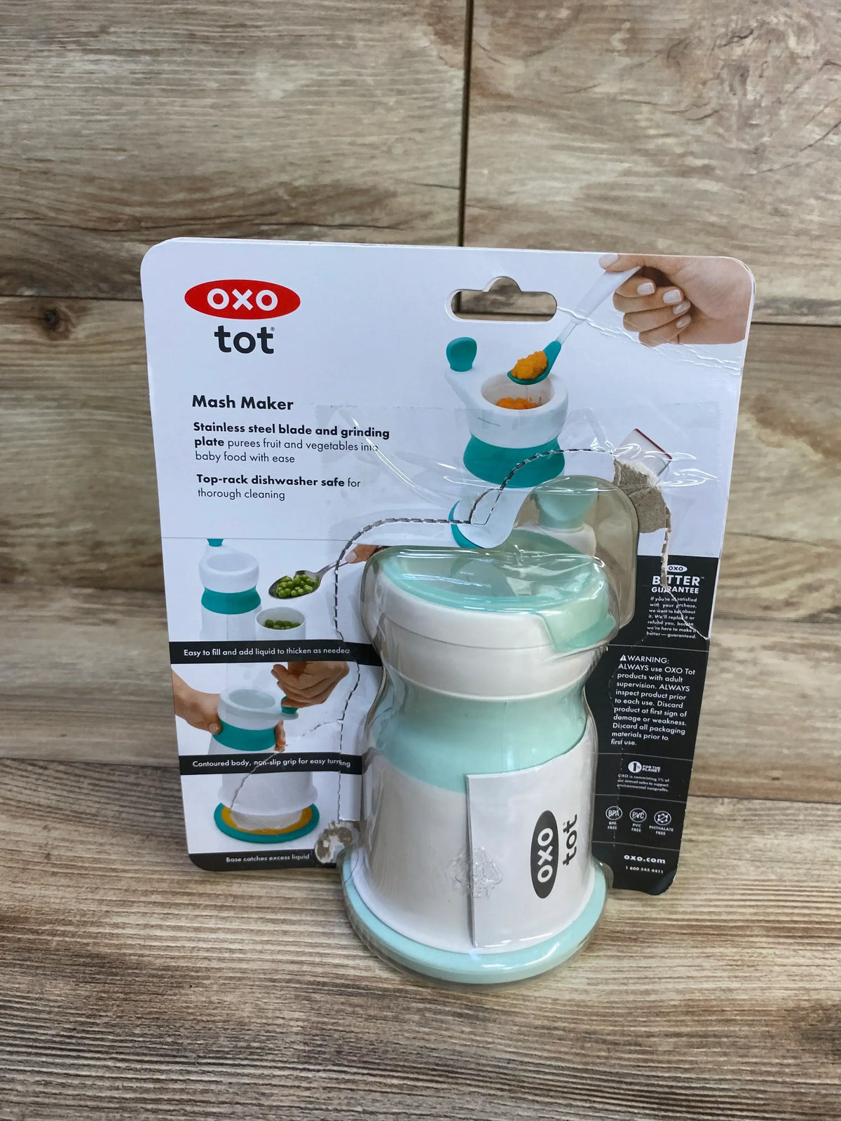 Front view of OXO Tot Mash Maker baby food mill in opal showing lid and suction base in original blister pack on wood table