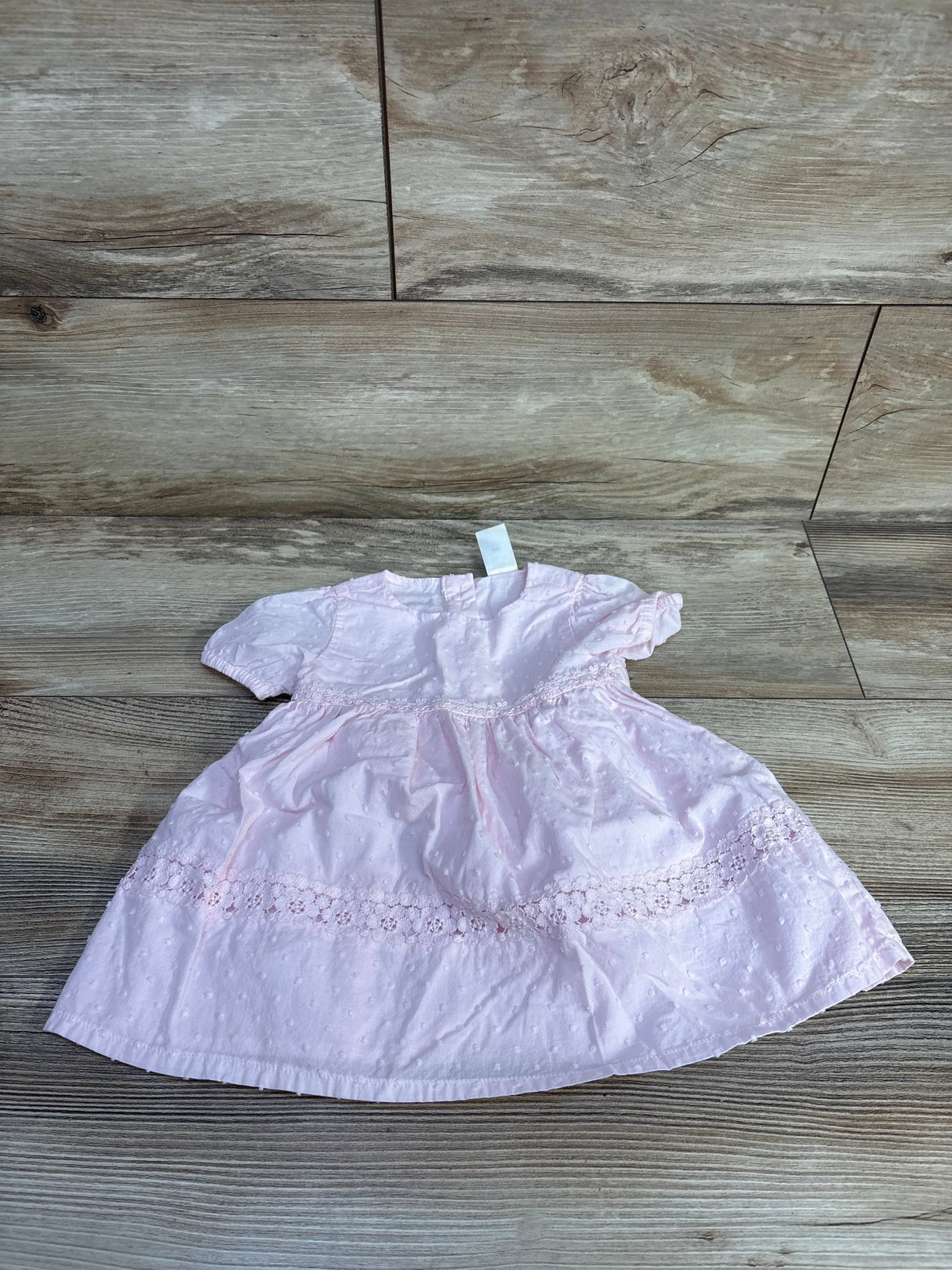 Front flat lay of OshKosh baby dress showcasing pink Swiss dot fabric and floral eyelet lace trim with wood plank background
