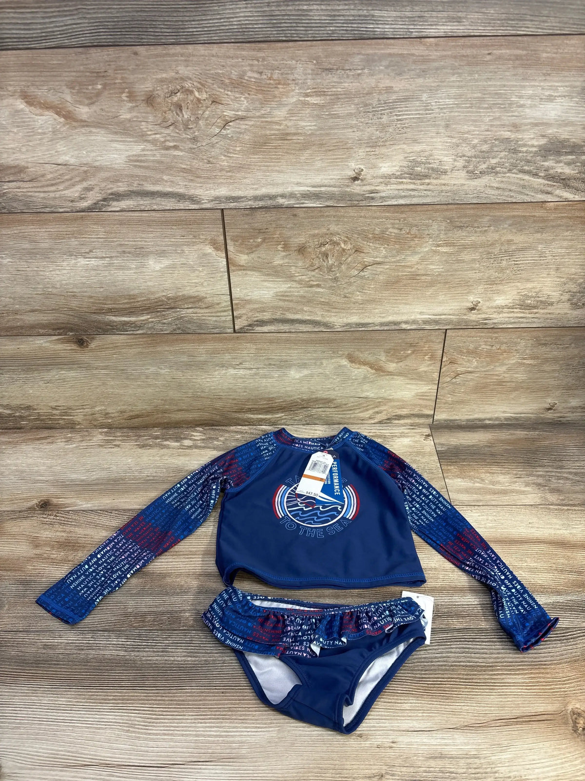 Flat lay of girls’ navy Nautica rashguard 2-piece set, long-sleeve top with wave graphic and ruffle bottoms on wood background