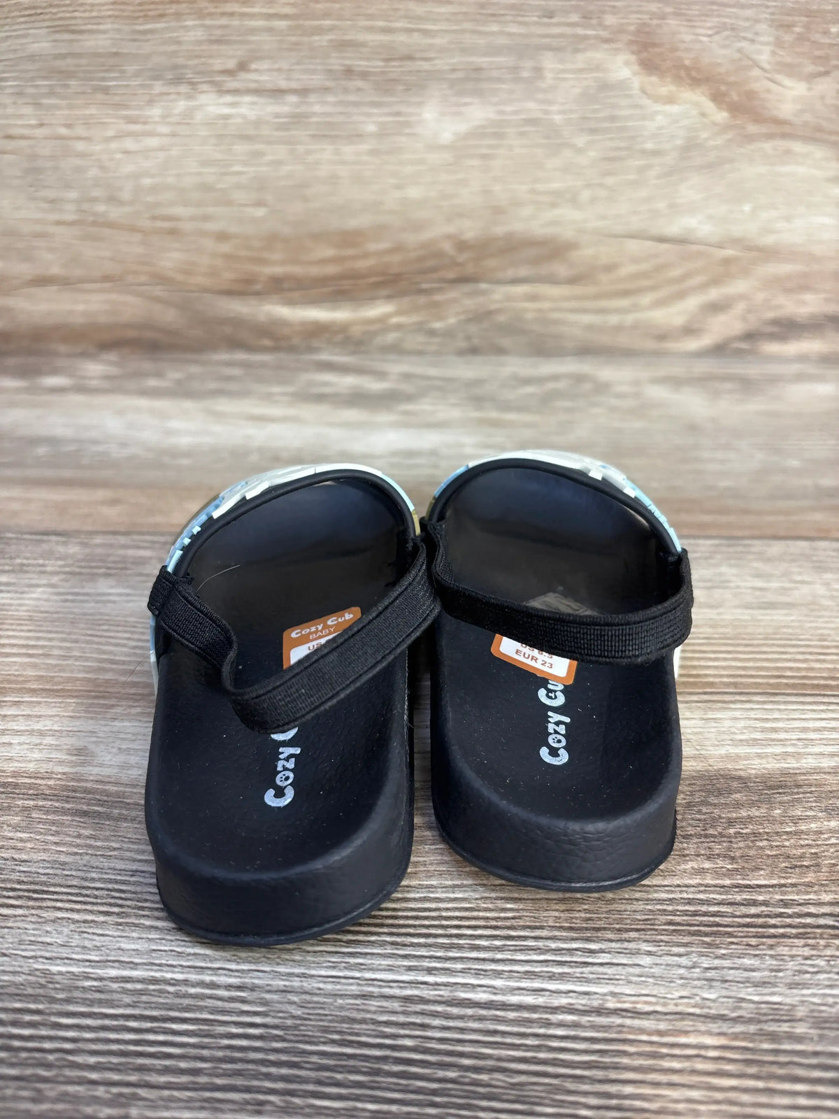 Pair of black Cozy Cub dinosaur slide sandals for kids on a wooden background.