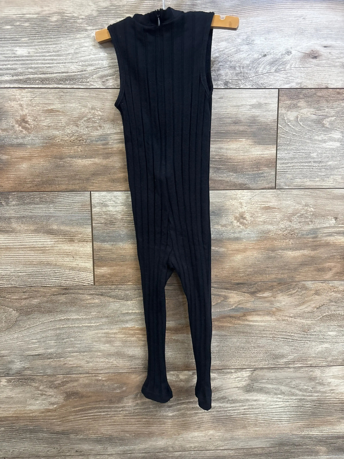 Front view of black ribbed toddler jumpsuit with footed legs on wooden hanger against wood plank background