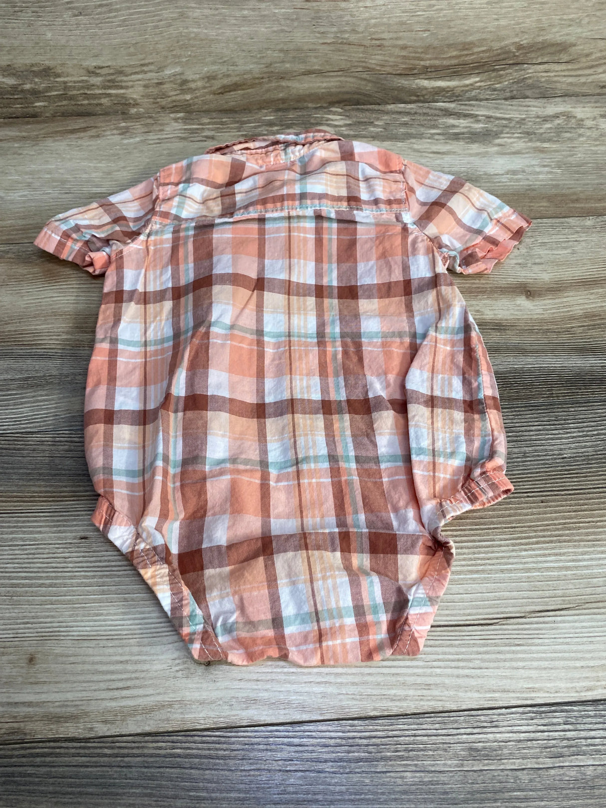 Back flat-lay of Oshkosh baby plaid button-up bodysuit in pink and peach showing short sleeves and elastic leg openings on a wood surface