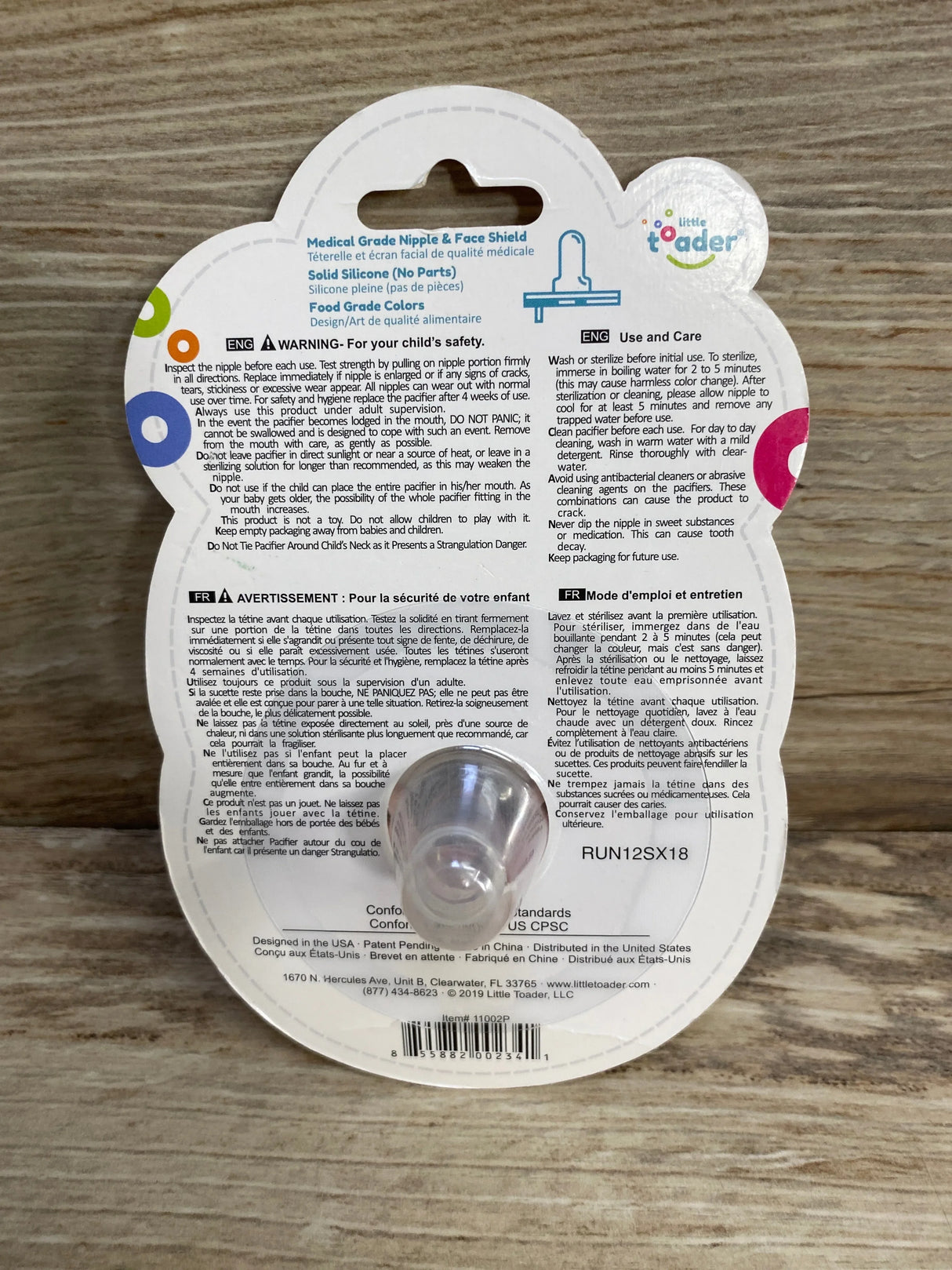 Front view of Little Toader Cupcake pacifier blister pack showing clear silicone nipple and printed care text on wooden table
