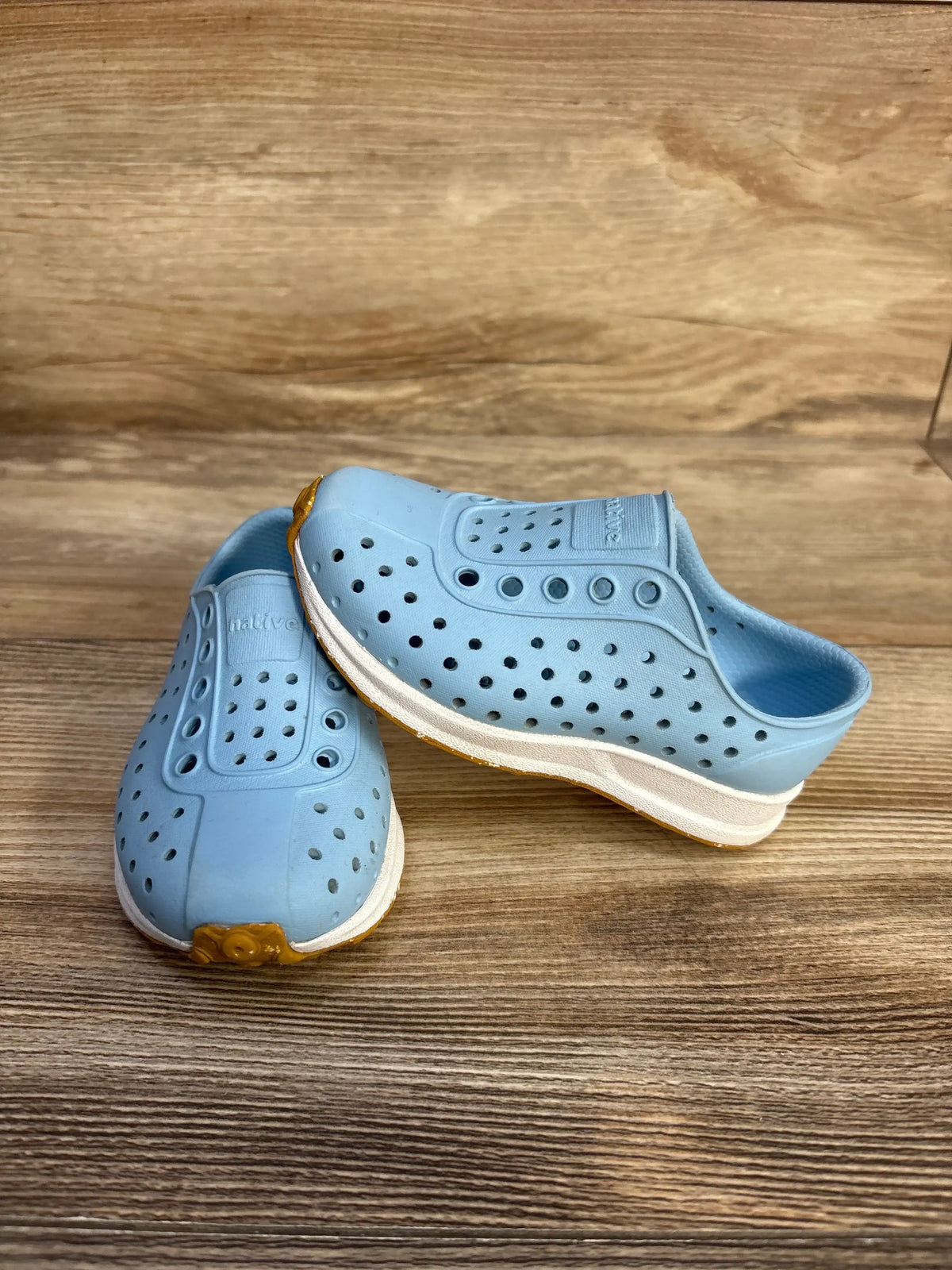 Angled view of blue Native Robbie Sugarlite slip-on shoes showcasing perforated uppers and white midsoles on wood