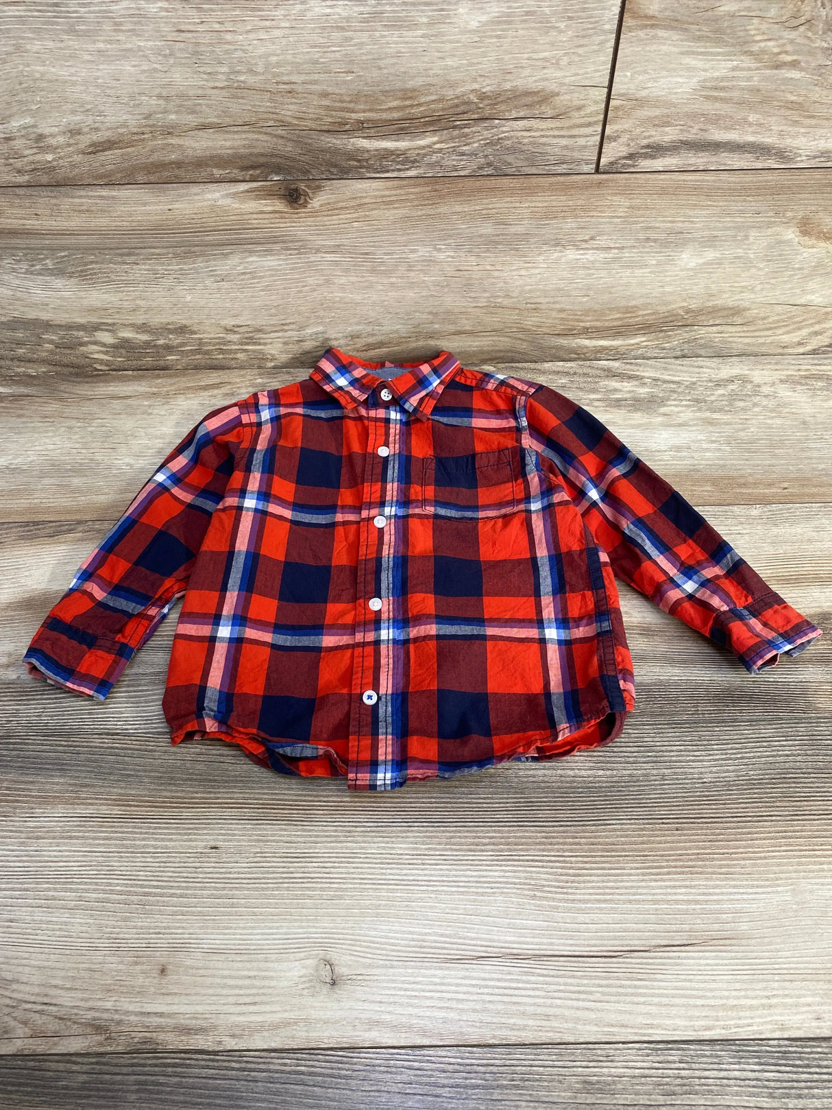 Front view of toddler red and navy plaid button-up shirt showcasing long sleeves, chest pocket, and white buttons on light wood background