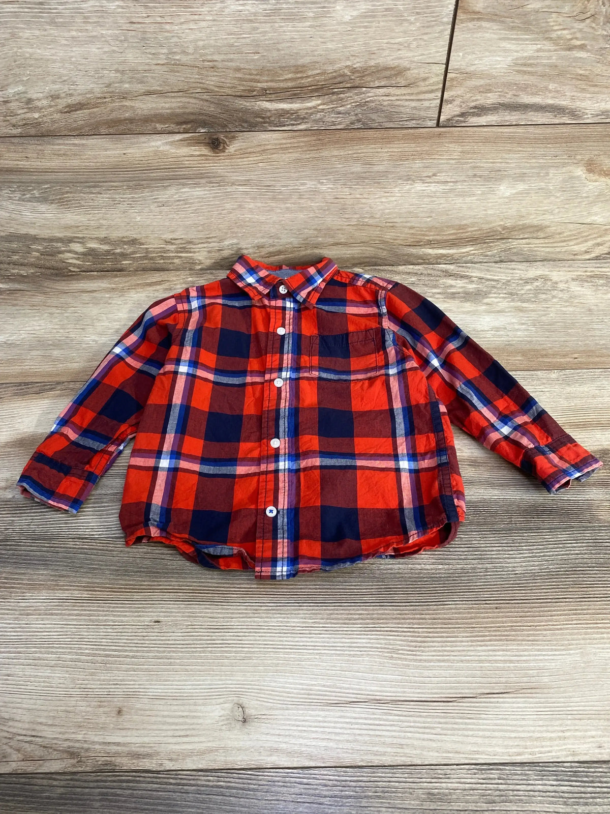 Front view of toddler red and navy plaid button-up shirt showcasing long sleeves, chest pocket, and white buttons on light wood background