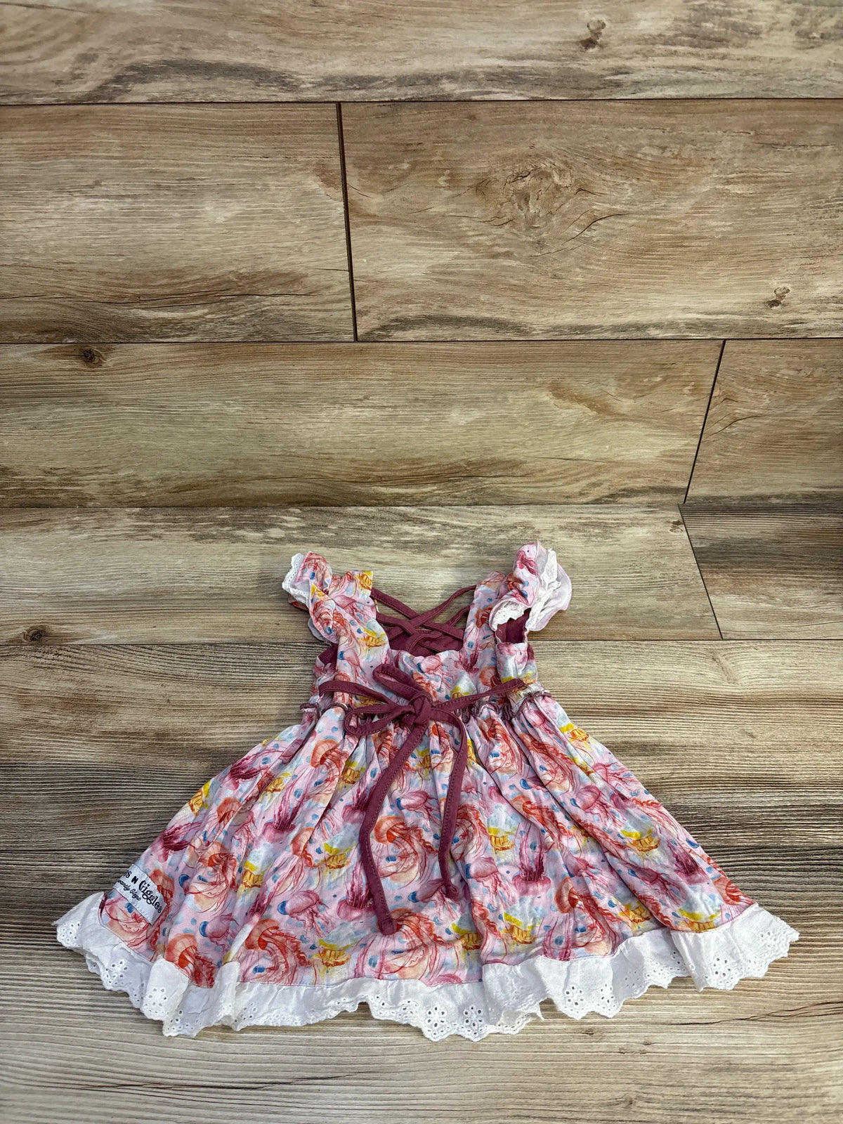 Top view of pink bamboo eyelet trim jellyfish dress showcasing printed jellyfish, white eyelet trim, and tie details on a wood-look floor