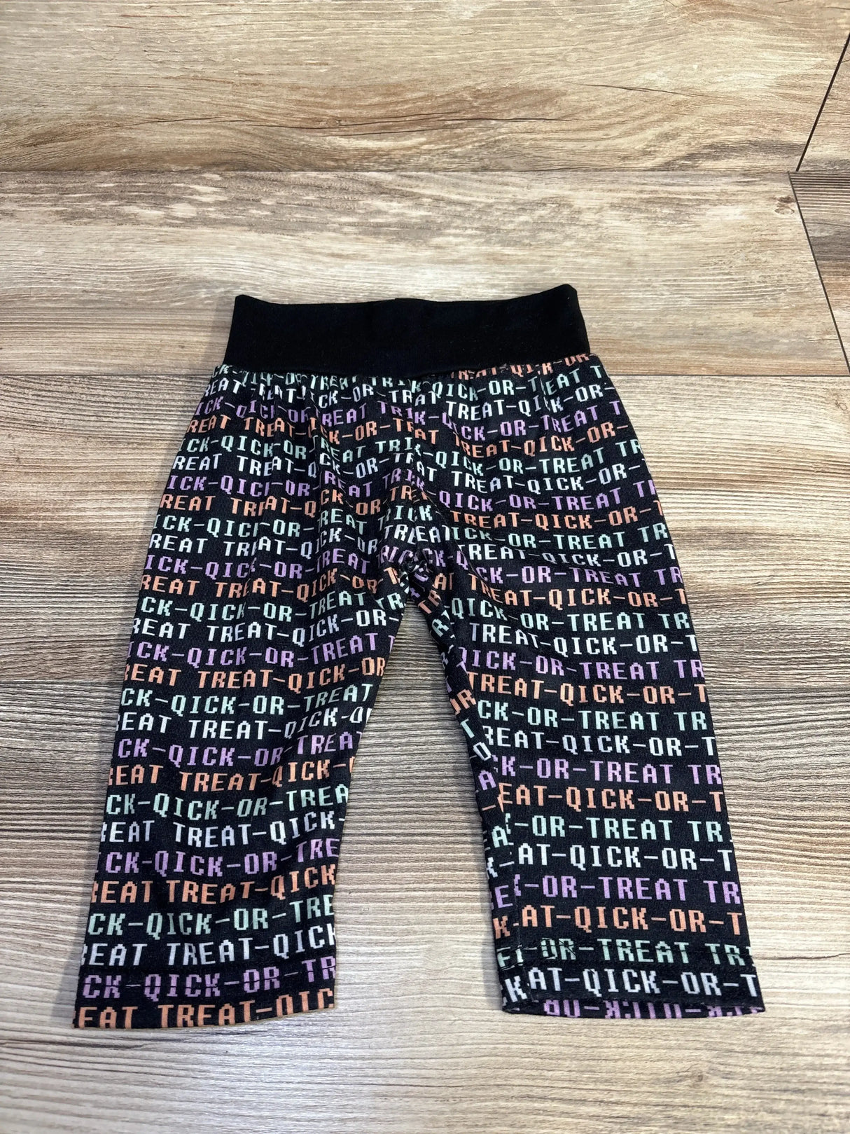 Flat lay of black Trick Or Treat pants in size 9 to 12 months with multicolored text pattern and elastic waistband on wood floor
