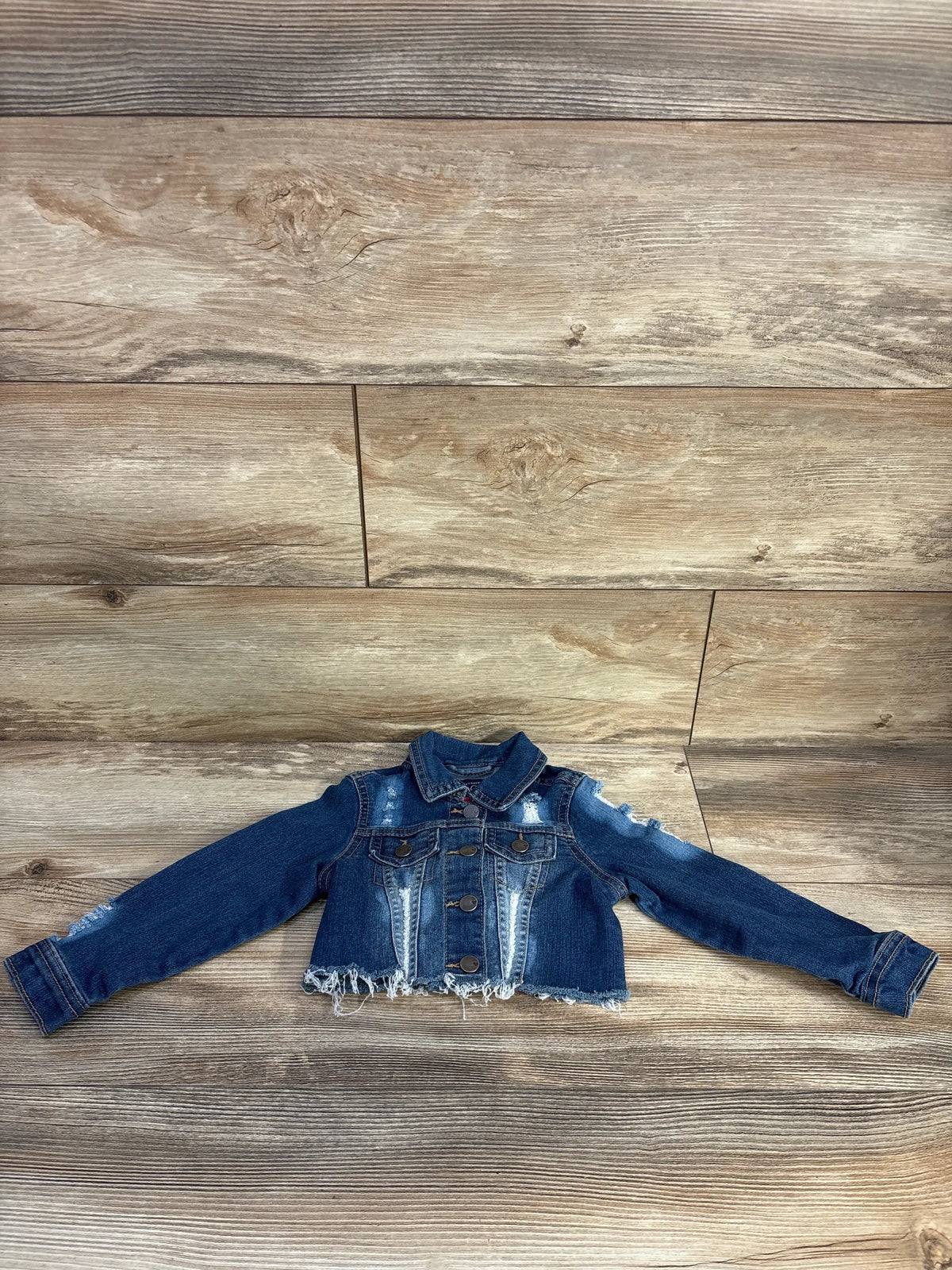 Front view of Children’s Place blue distressed denim jacket in size 2T showcasing frayed detailing, laid flat on wood